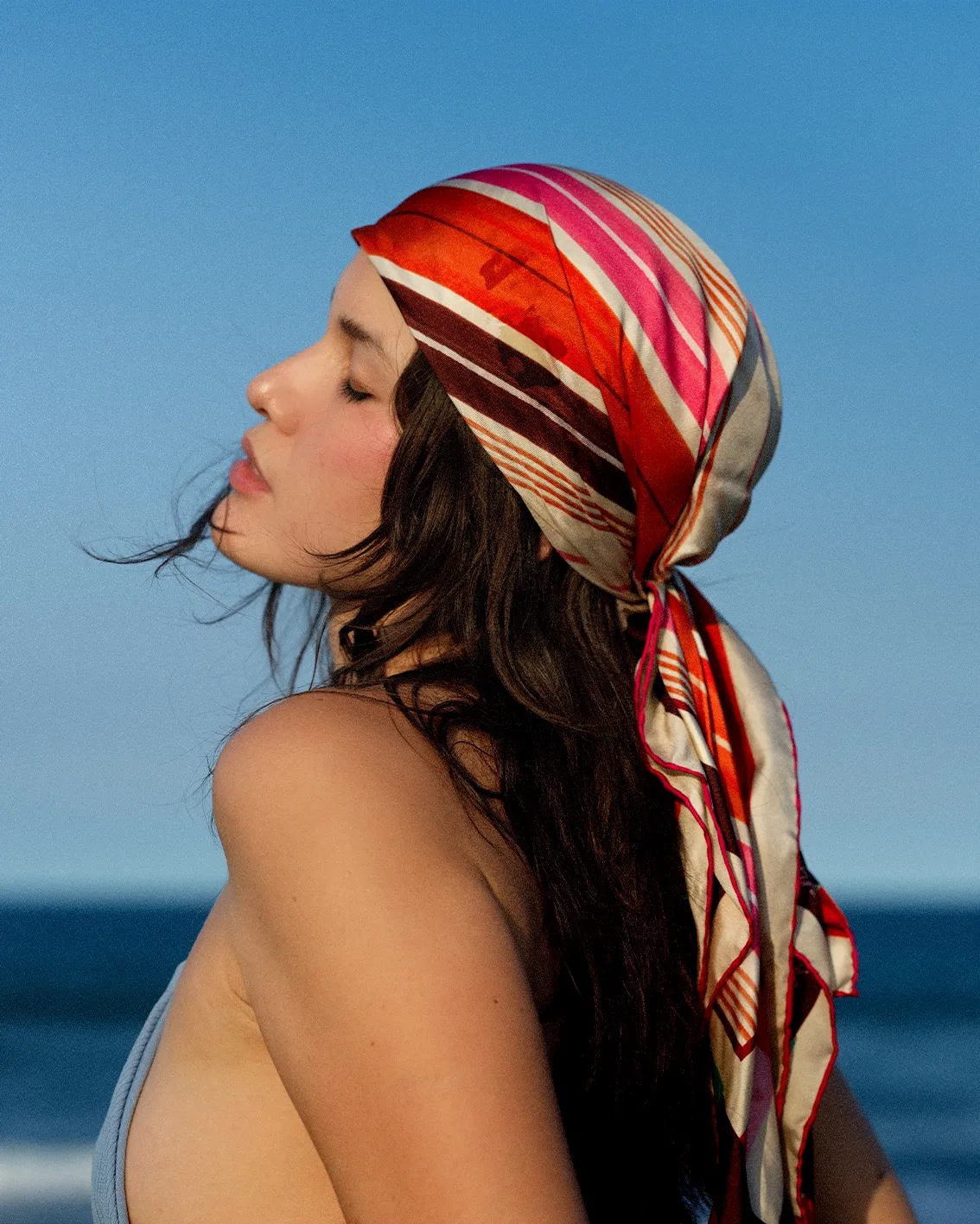 Profile view of a woman with a colorful striped headscarf tied at the back, against a blue sky and sea.