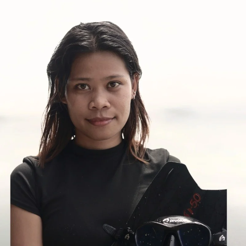 Portrait of a young woman in a black shirt standing outdoors by a calm body of water, with a black wakeboard in front of her.