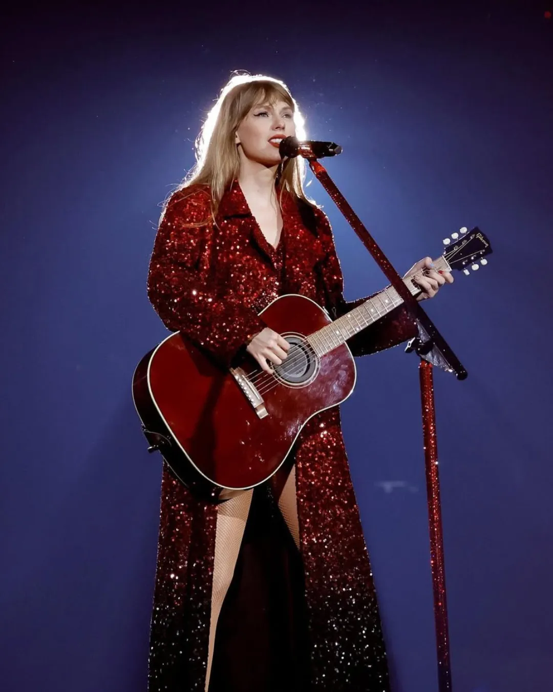 Singer performing on stage with a red sequined gown, playing a red acoustic guitar in front of a microphone.