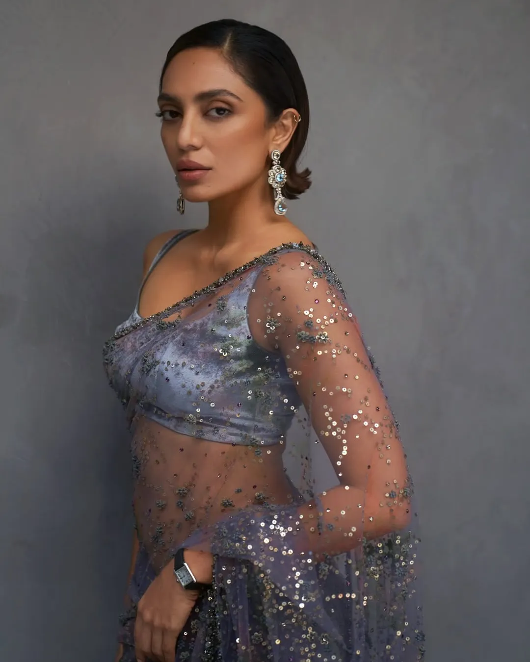 Portrait of a woman wearing a sequined lavender saree with sheer embellished sleeves, posing against a gray backdrop, with chandelier earrings.