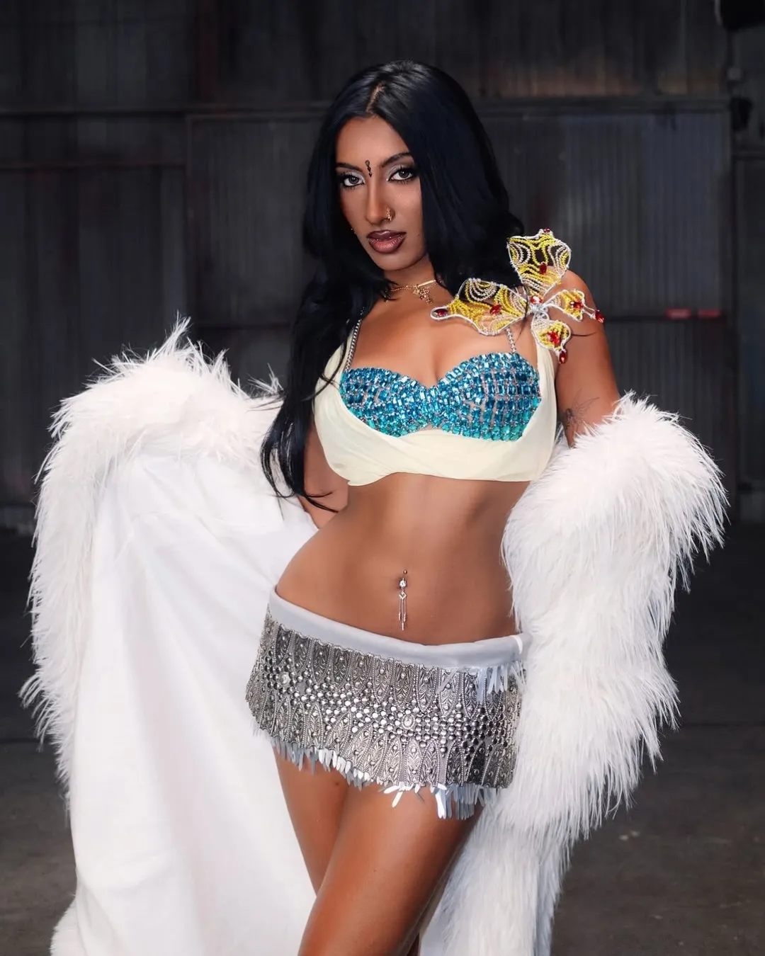 Model in a turquoise beaded bustier, white fur coat, and a silver fringe mini skirt posing in a dark industrial setting.