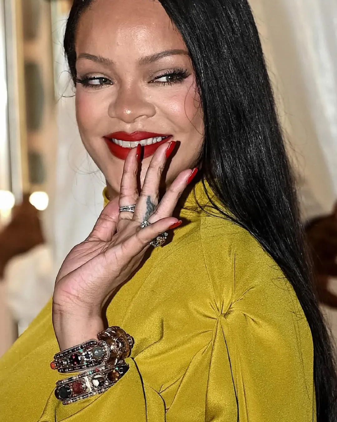 Smiling woman with long black hair, wearing a mustard-yellow top, bold red lipstick and multiple bracelets on her wrist.