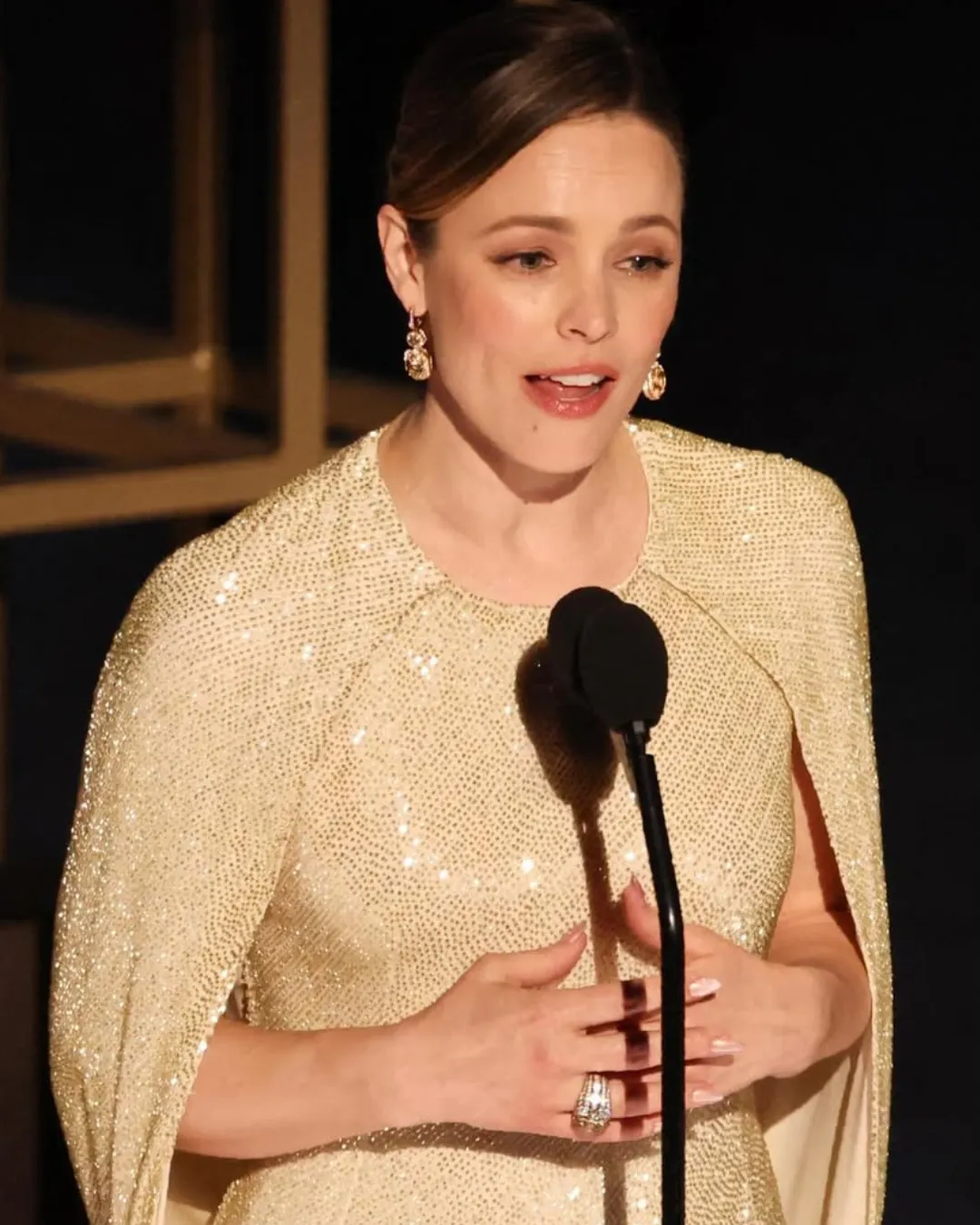 A woman in a glittering gold dress speaks into a microphone, hands held at chest, wearing dangling gold earrings.
