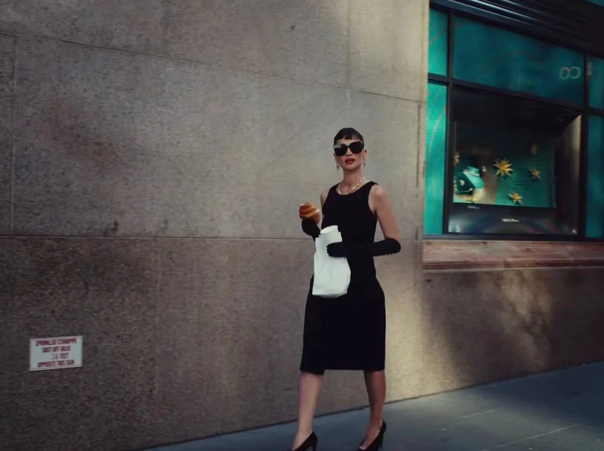 Woman in a black sleeveless dress with long gloves and sunglasses, holding a white bag and a pastry on a city sidewalk.