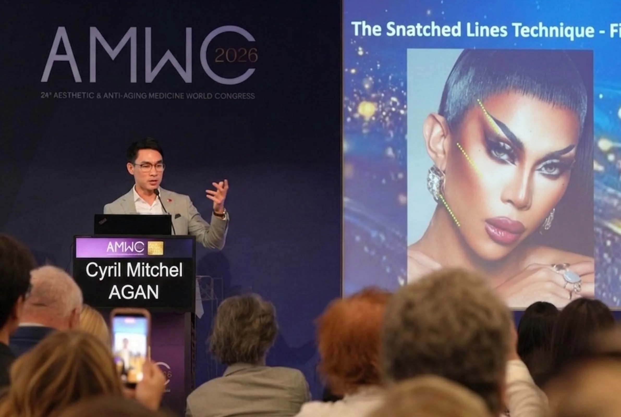 Speaker at a podium on an AMWC 2026 stage; large banner and a giant portrait backdrop visible on the right.