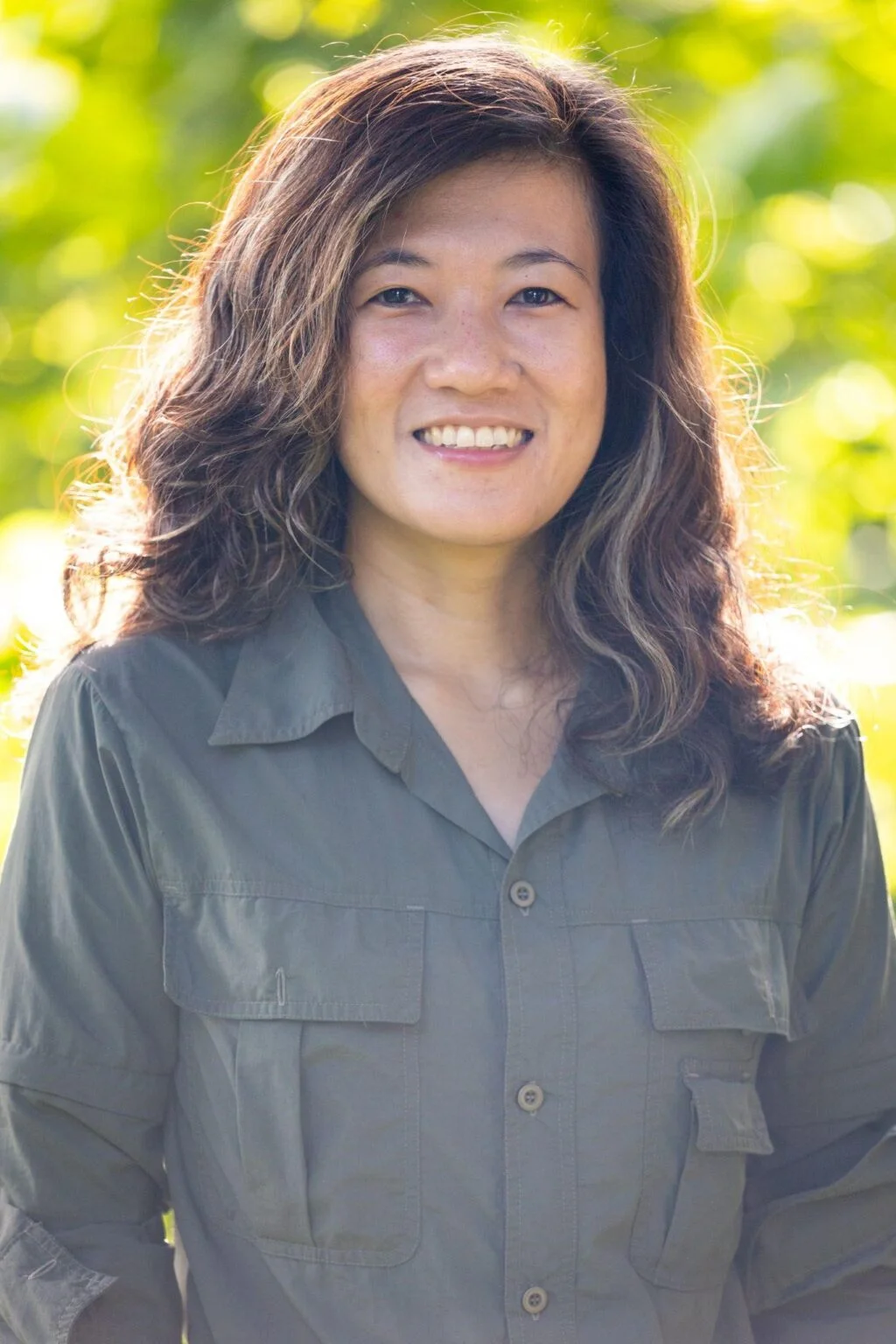 Smiling woman with wavy hair outdoors in sunlight wearing a gray button-up shirt.