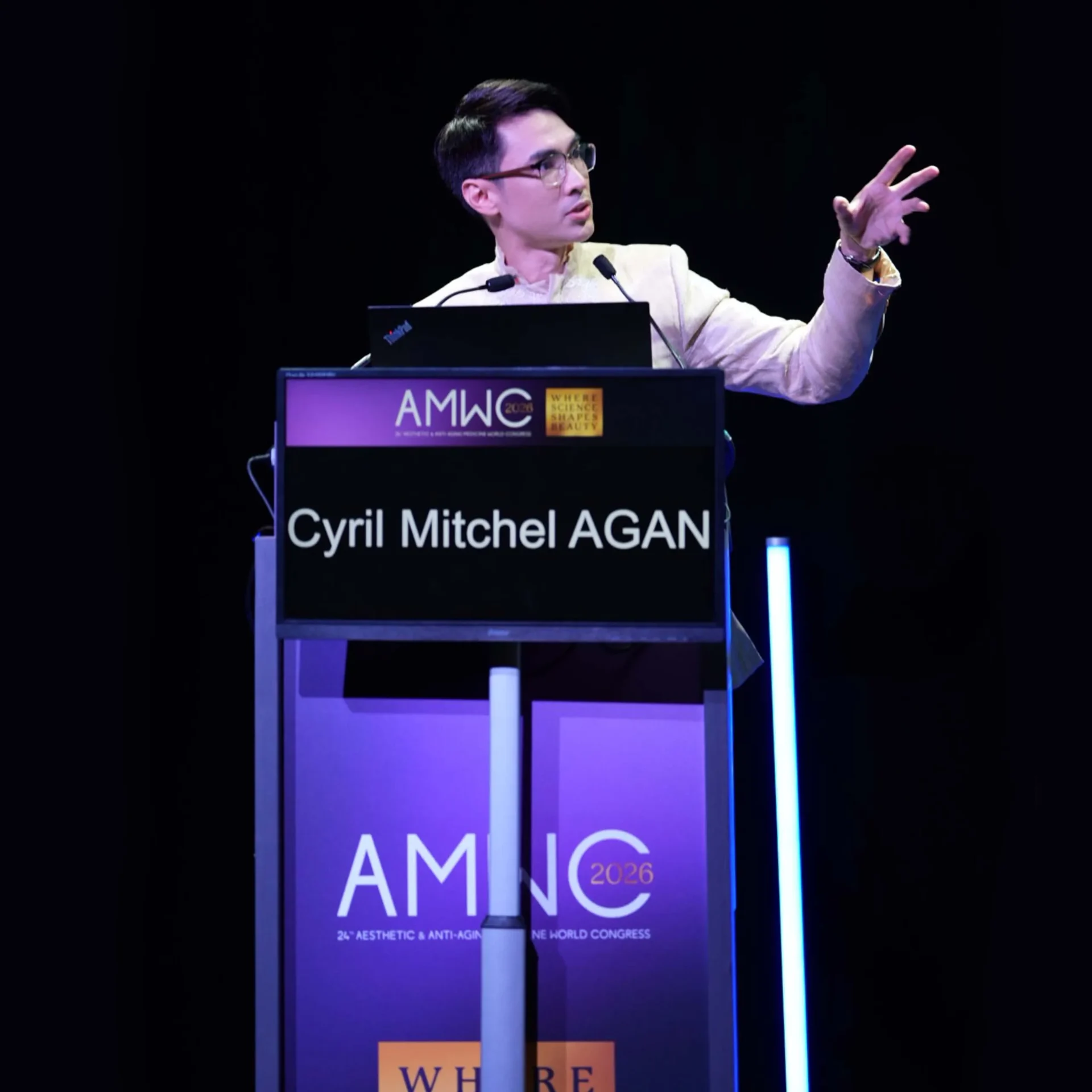 Male speaker at a podium on a dark stage, gesturing with his right hand; banner shows AMWC 2026 and 'Where science shapes beauty' text.