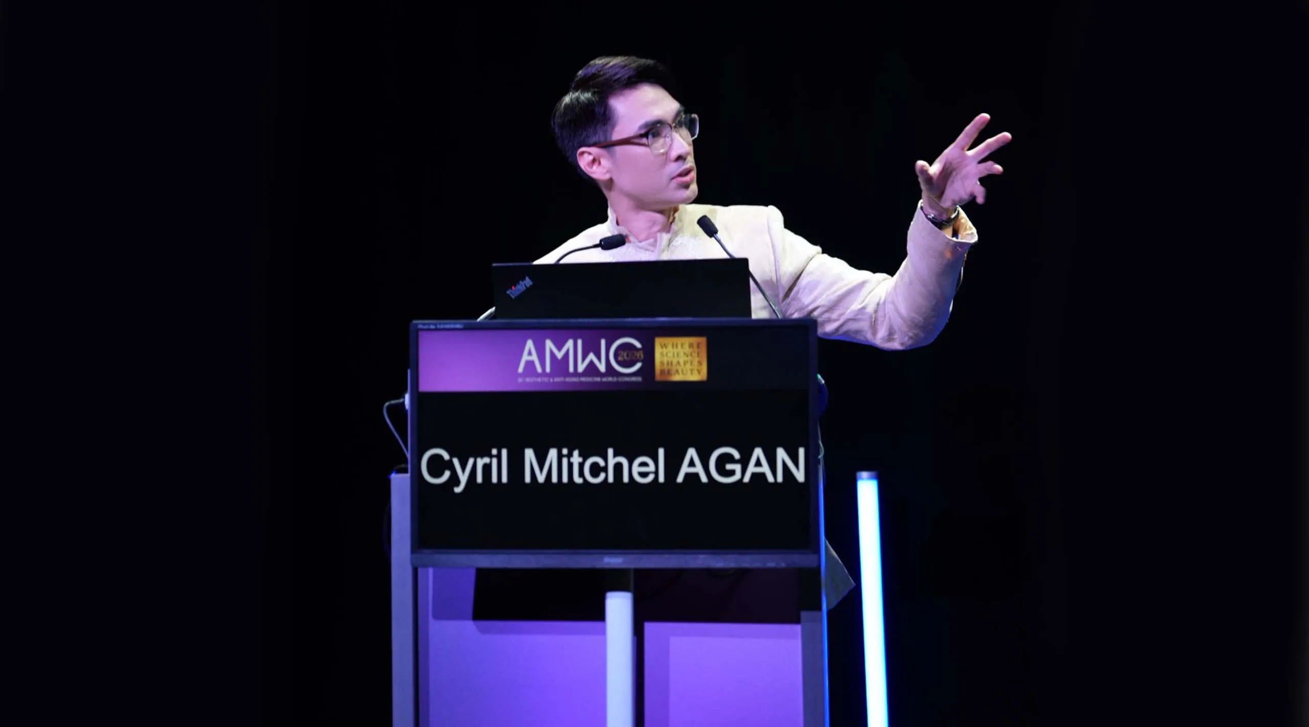 Speaker at a podium delivering a talk, gesturing with his right hand toward the audience; podium signage reads Cyril Mitchel AGAN.
