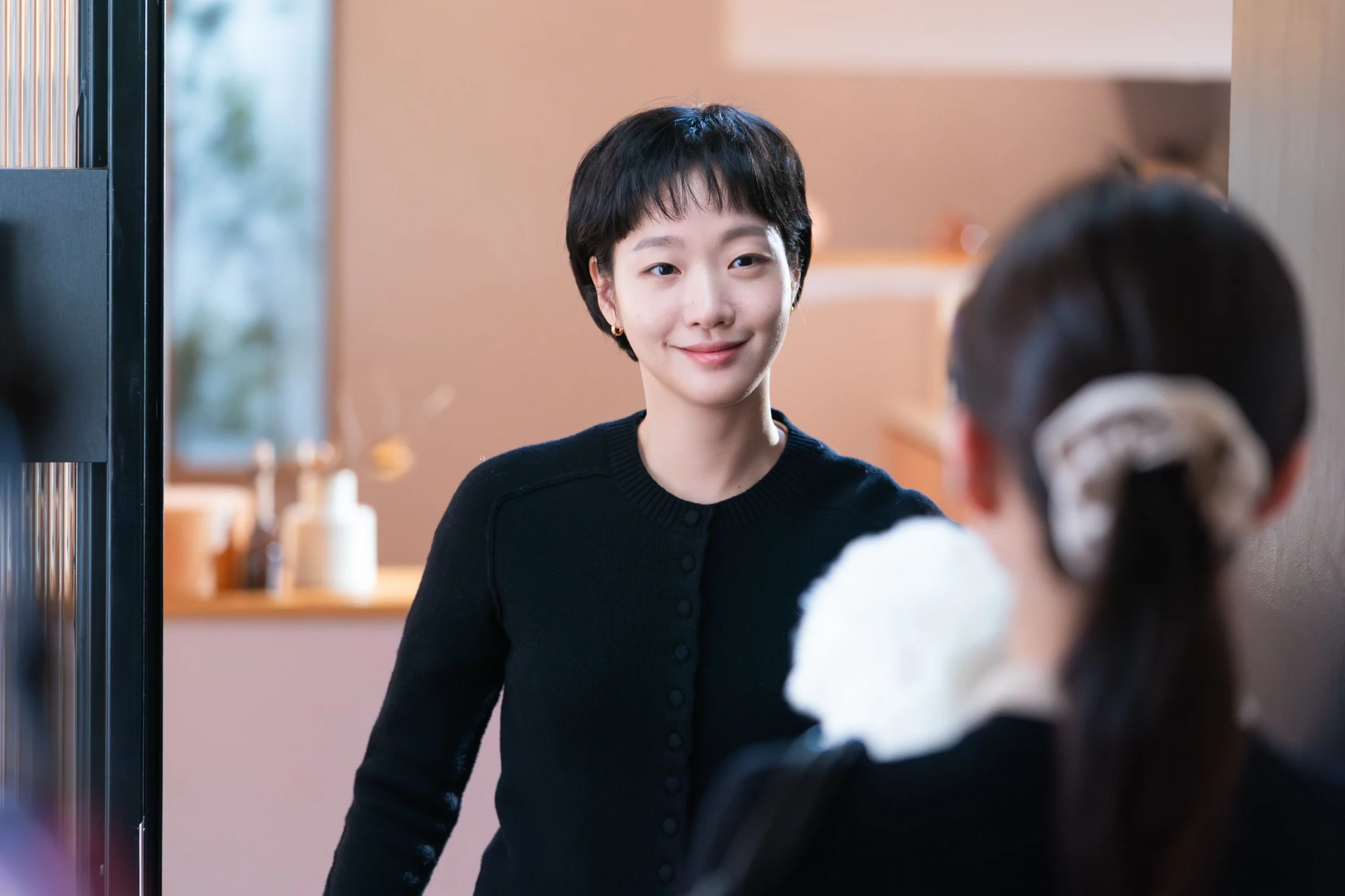 Woman with short black hair smiling at a person off-camera inside a store, wearing a black cardigan against a warm background interior.