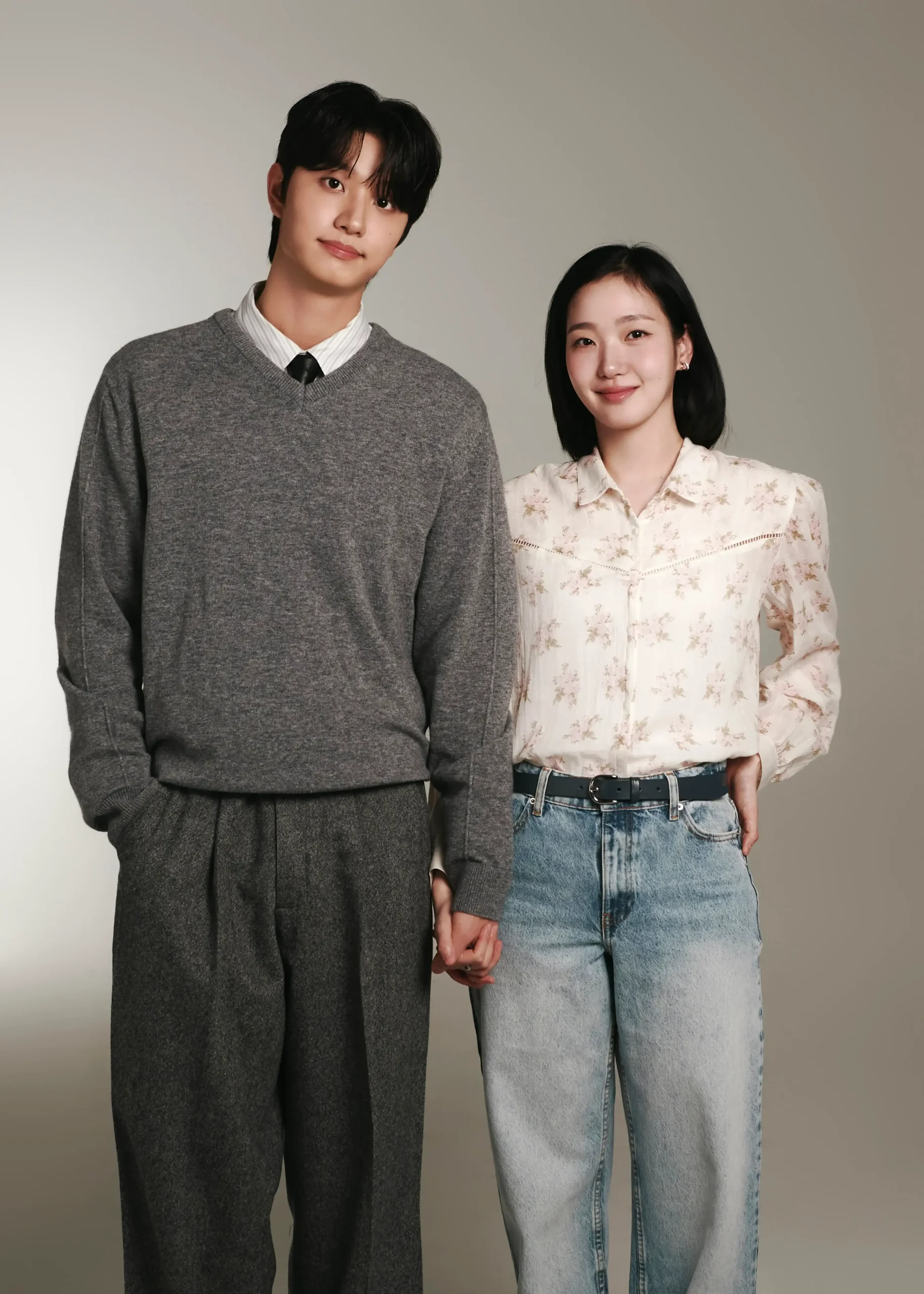Young man in a gray knit sweater stands beside a woman in a cream floral blouse and jeans; they are holding hands in a studio portrait.