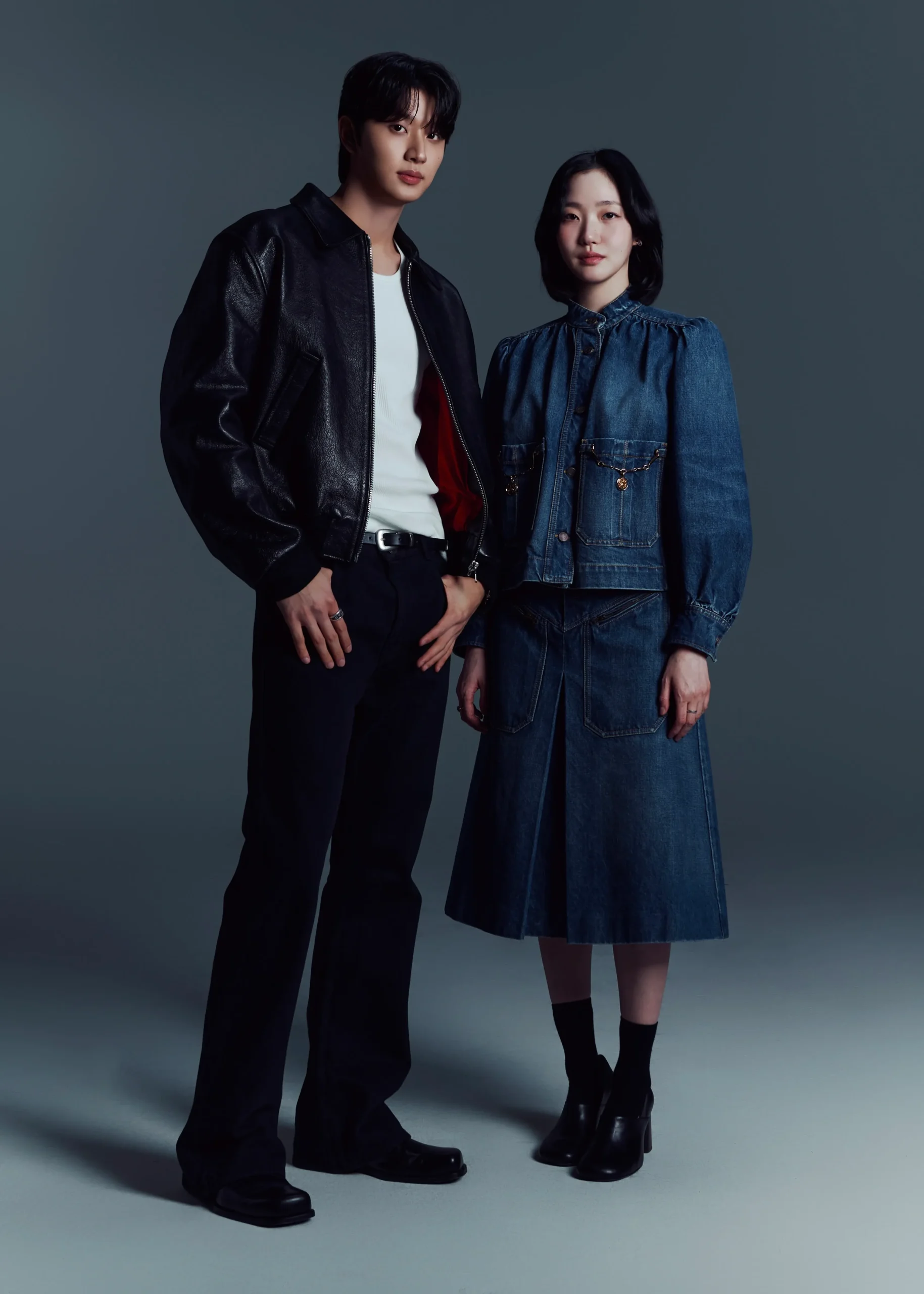 Two fashion models pose side by side in a studio: man in a black leather jacket and white tee, woman in a denim dress with pockets and a chain detail against a gray backdrop.