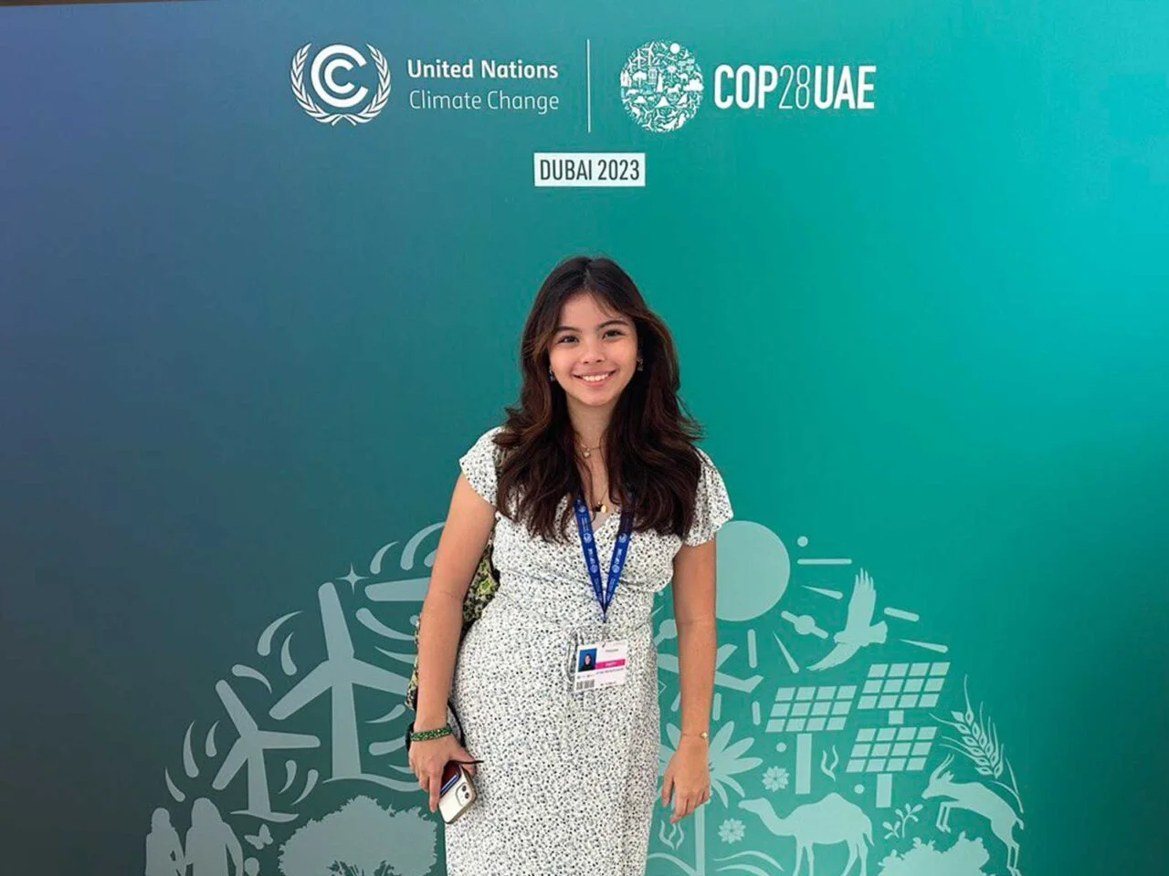 Smiling woman with lanyard posing in front of a COP28 UAE Dubai 2023 backdrop at a climate conference.