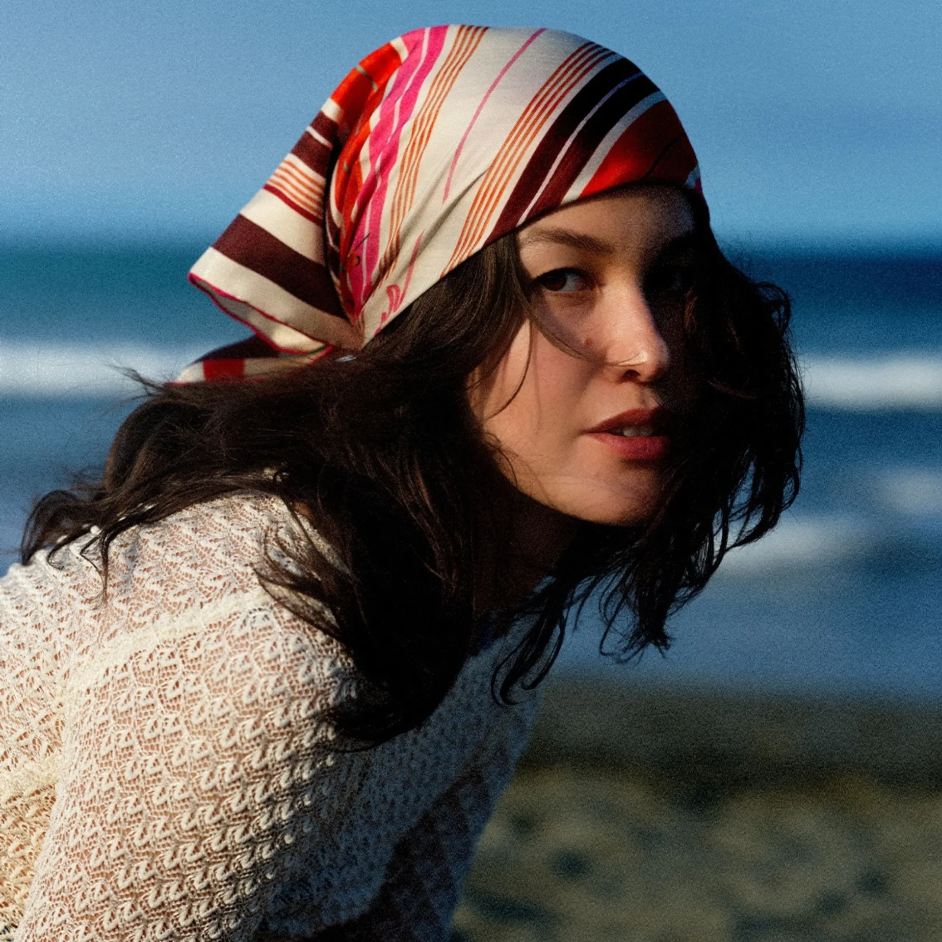 Woman at the beach wearing a striped headscarf and white lace top, looking over her shoulder toward the camera.