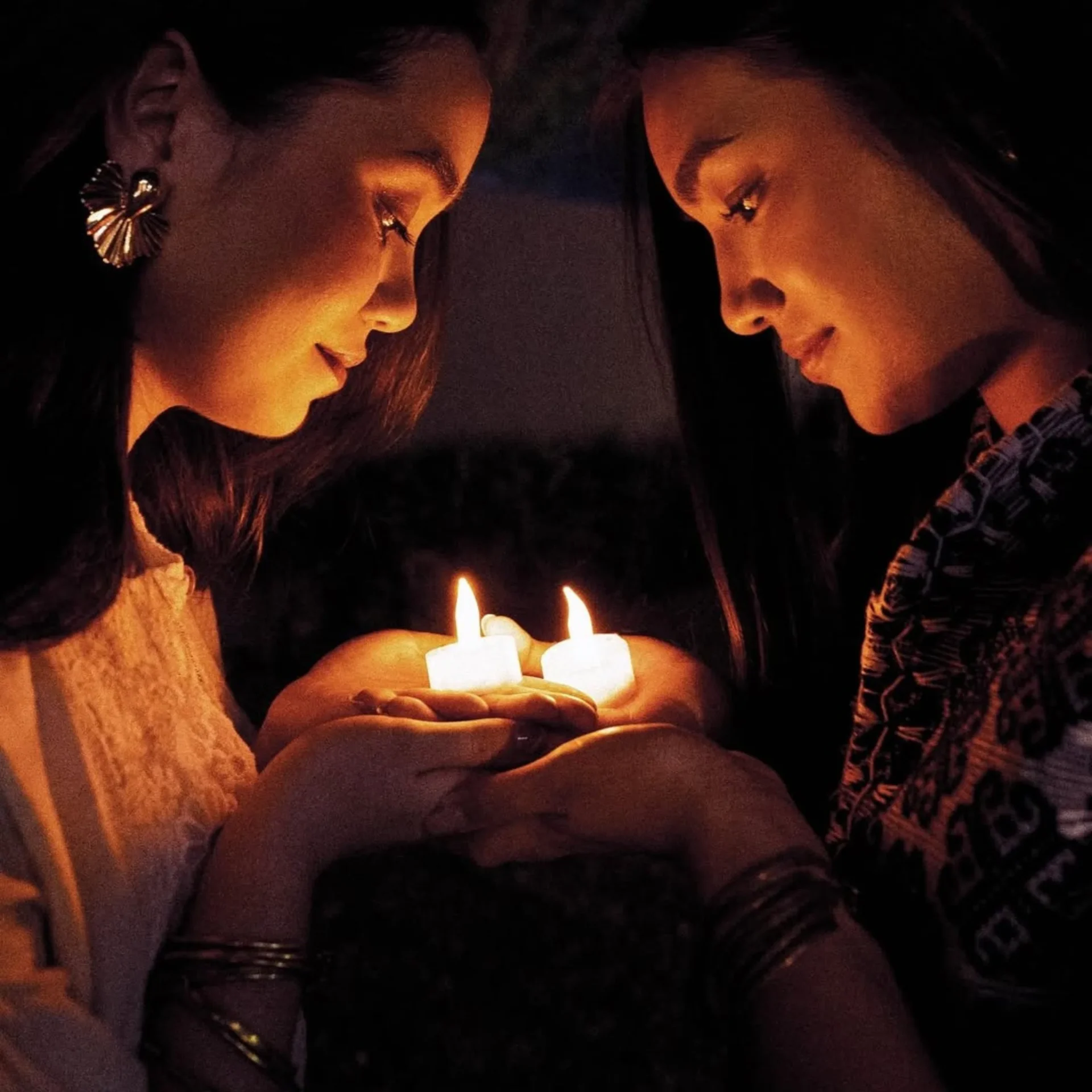Two women share a candlelit moment, cupping the flames in their hands and smiling softly at each other.