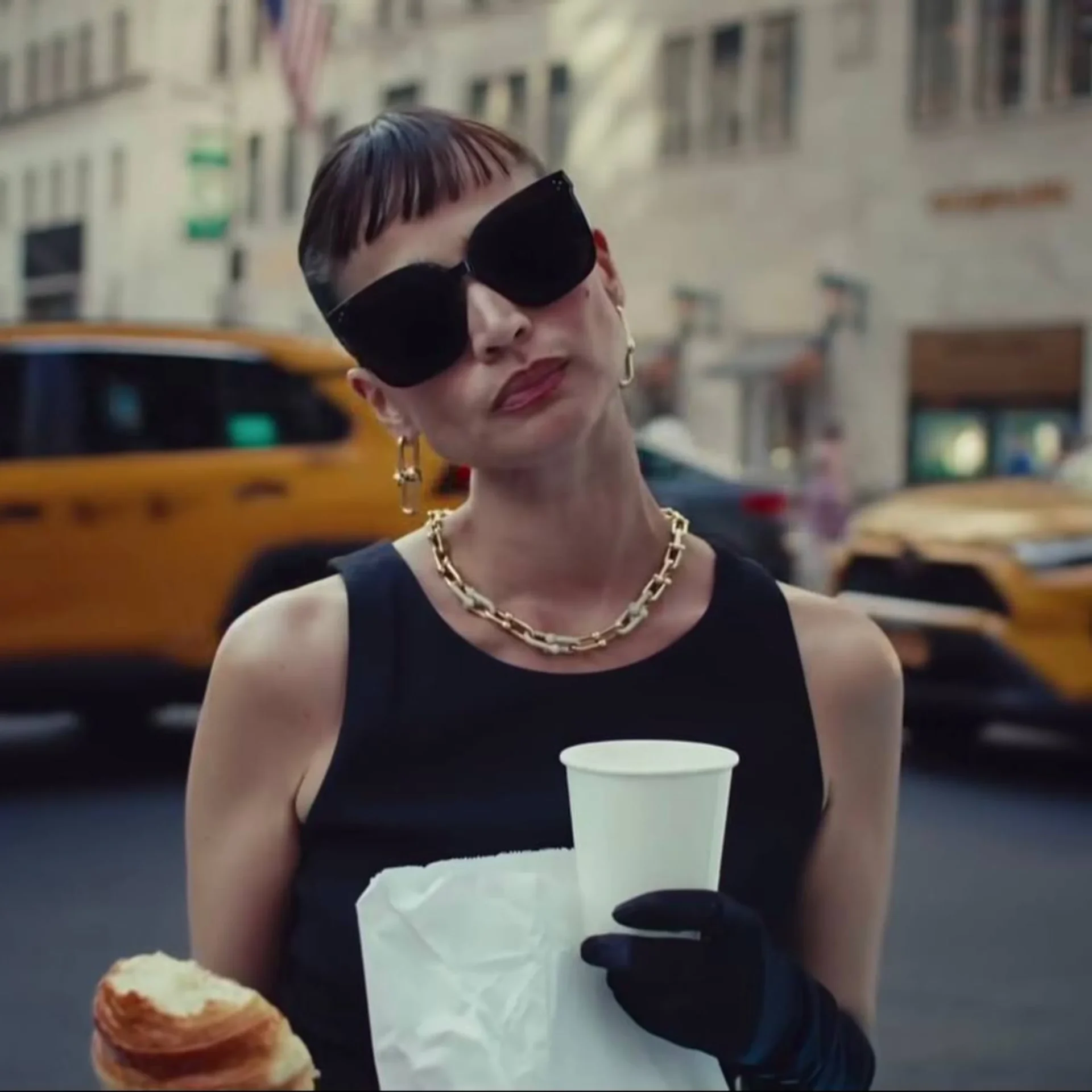 Portrait of a stylish woman in a city street, wearing oversized black sunglasses and gold jewelry, holding a white cup and a pastry in her hands.