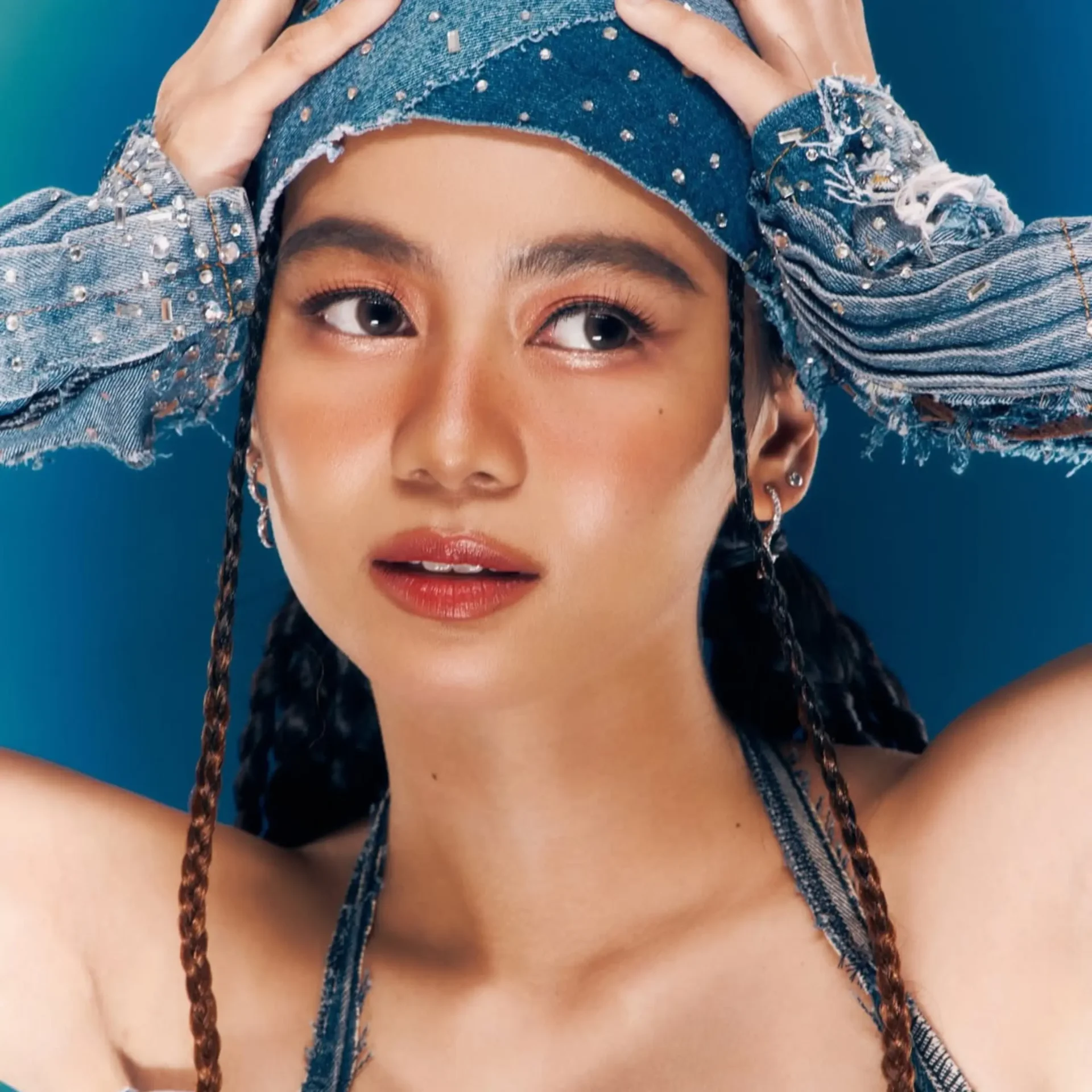 Close-up portrait of a woman with denim jacket and beaded details, hands on her head against a blue background, eyes looking to the side.