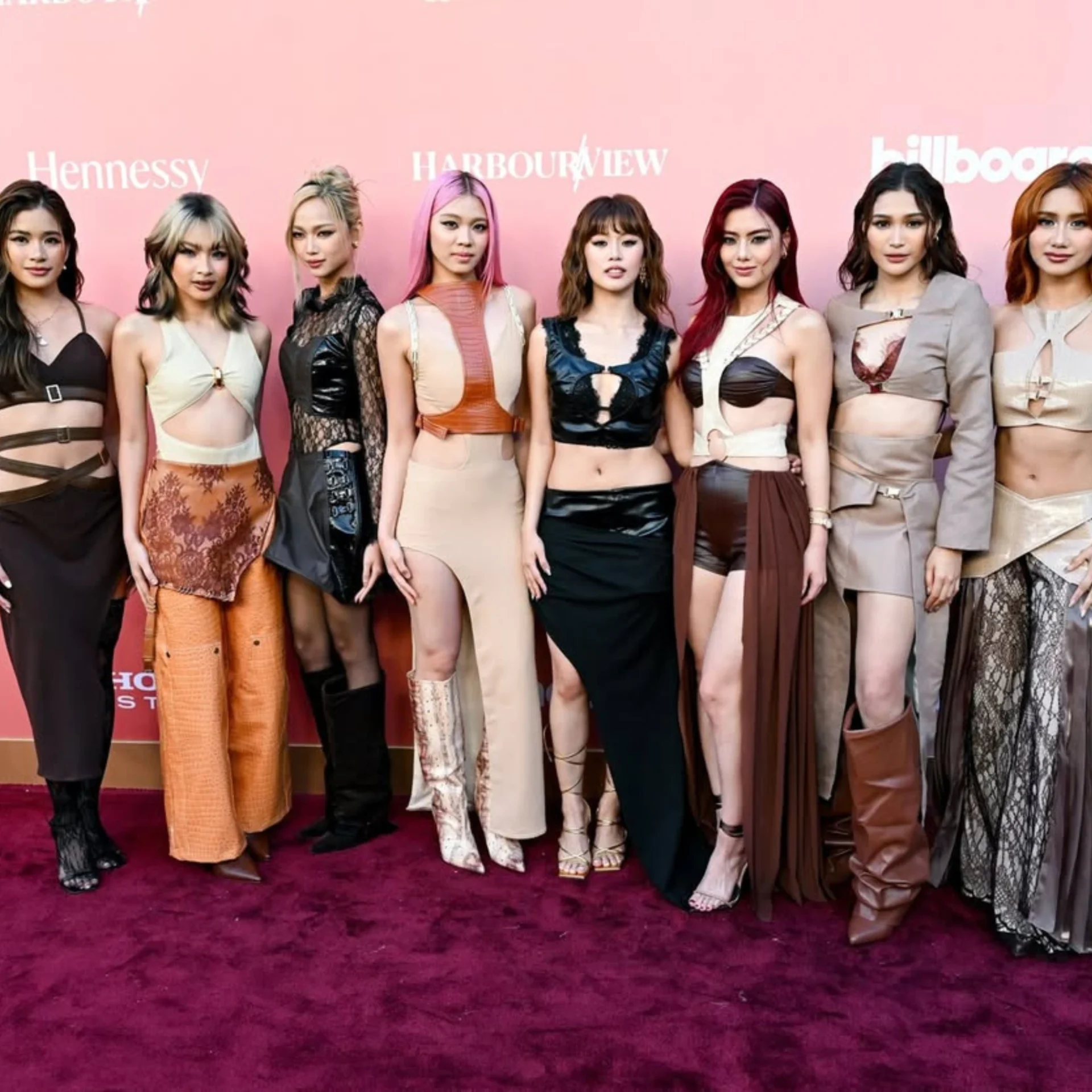 Nine models posing side-by-side on a red carpet in front of a pink backdrop at a fashion event, wearing varied outfits and high heels.