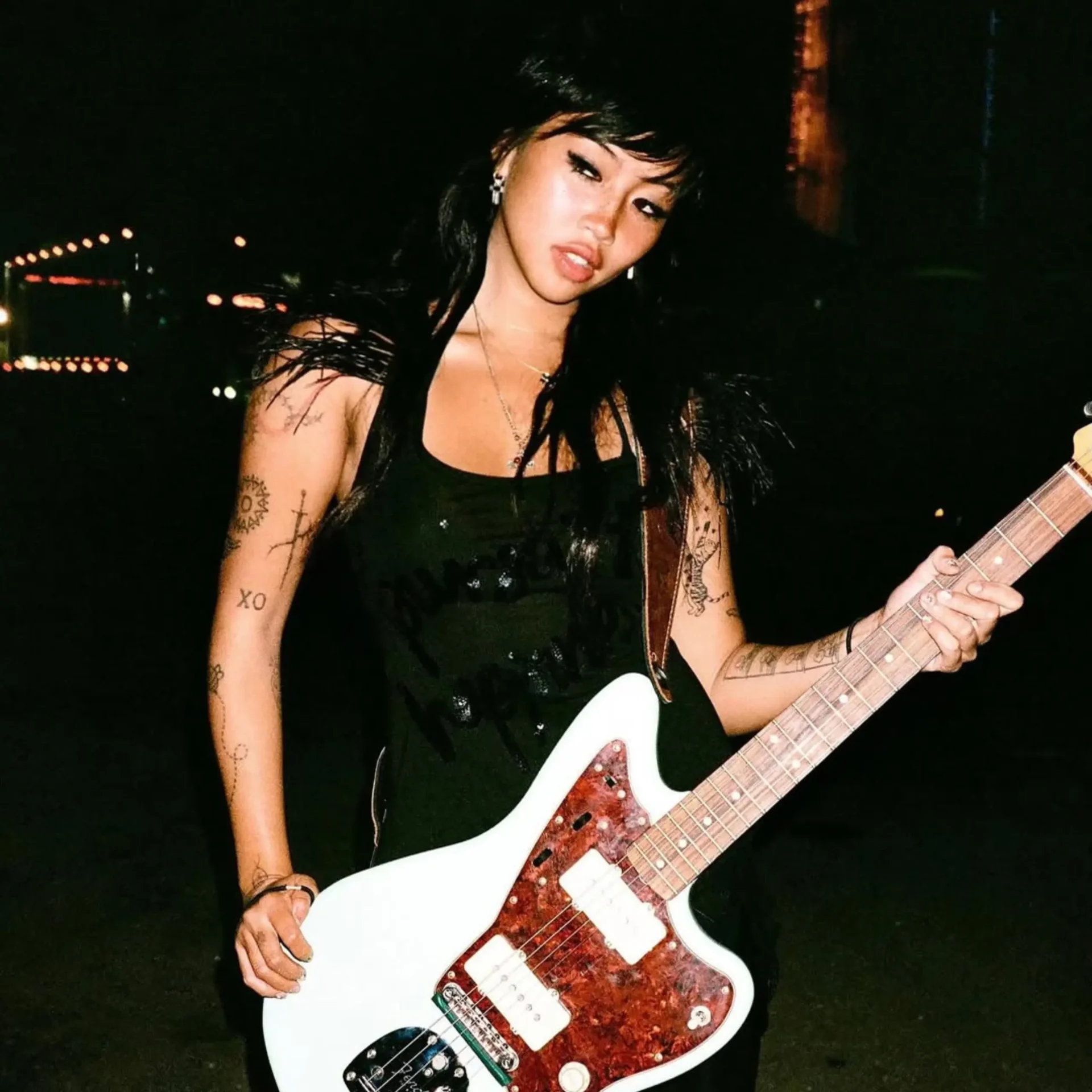 Woman with dark hair in a black outfit playing a white electric guitar at night, tattoos visible on her arms.