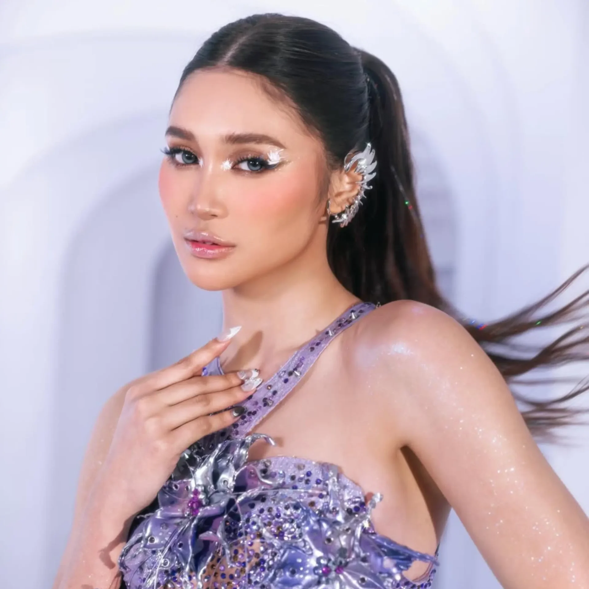 Close-up of a woman with blue eyes, bold glitter eye makeup, and a lavender sequined dress; hair in a ponytail with a silver hair accessory.