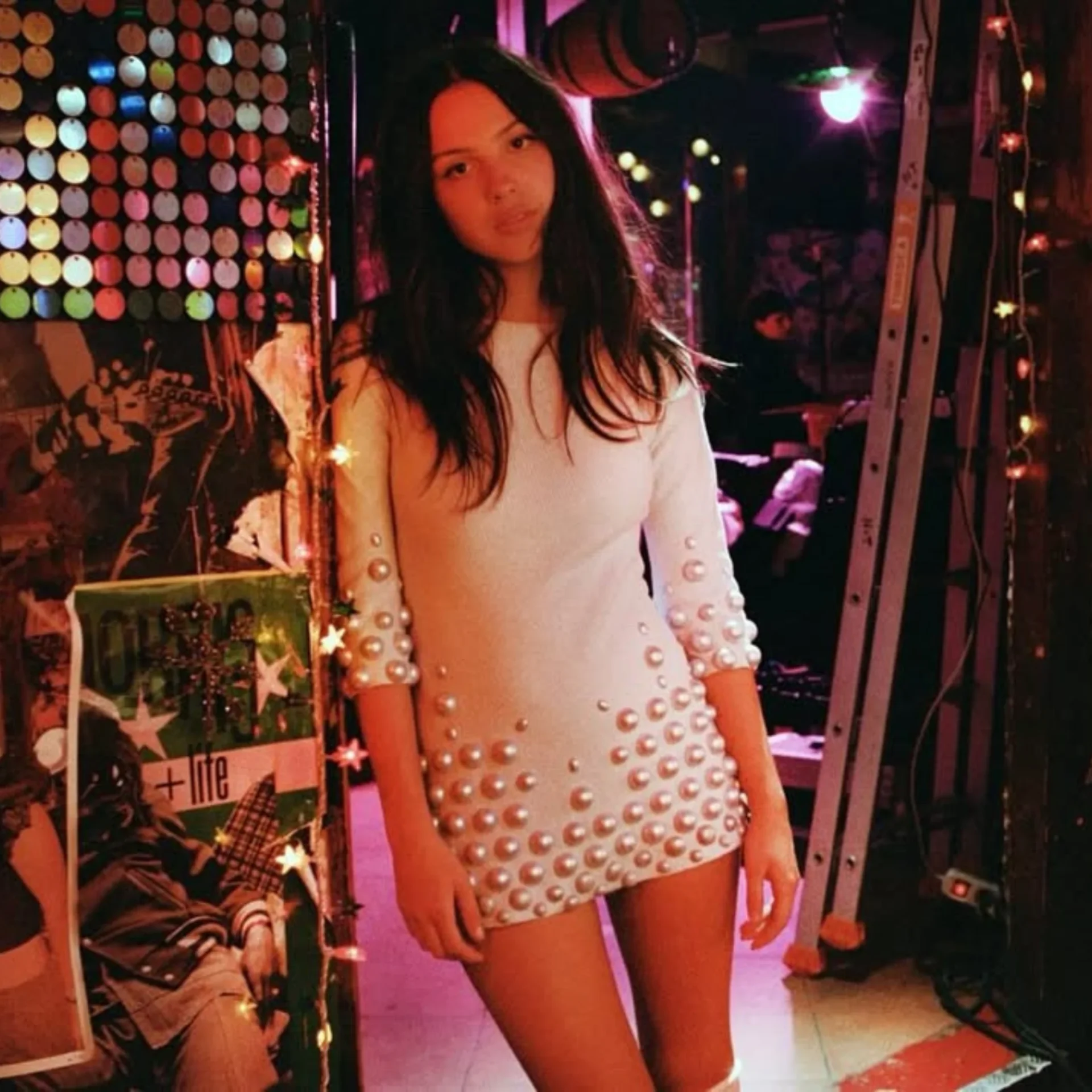 Young woman in a white pearl-embellished mini dress posing in a backstage area with colorful lights and string lights nearby.