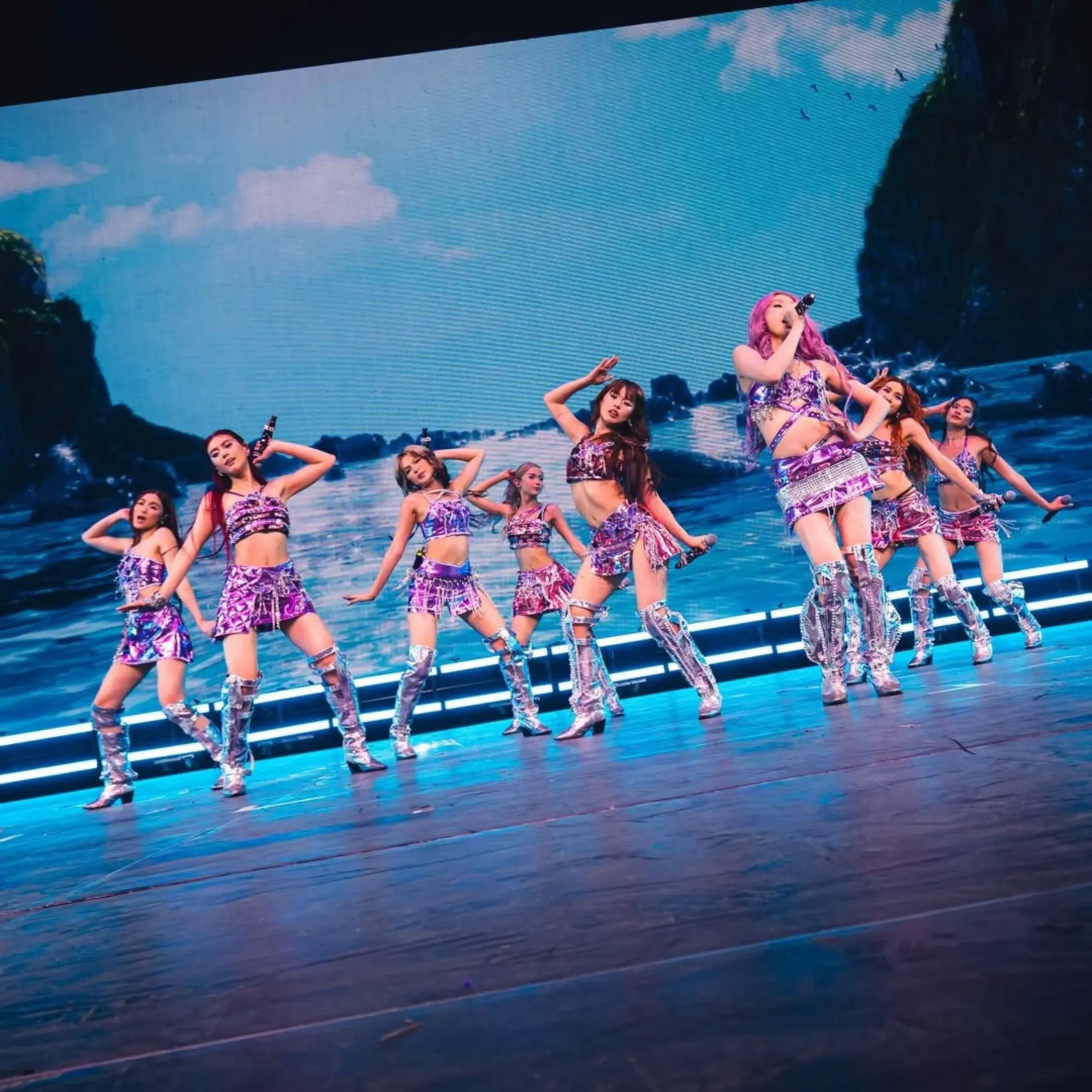 Pop music performance on a blue-lit stage: lead singer with pink hair holding a microphone, surrounded by dancers in matching purple outfits.