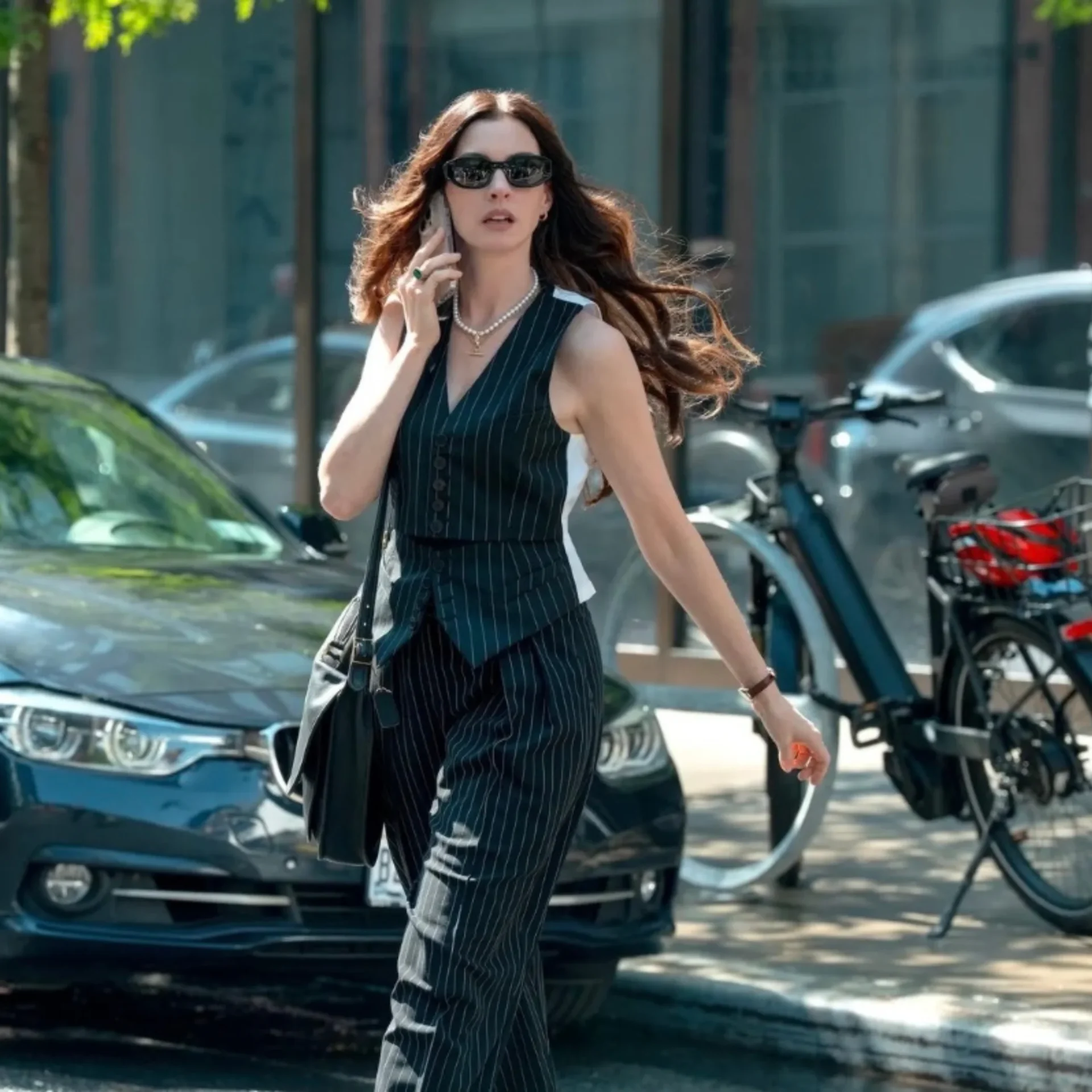 Woman in a pinstripe dress talking on her phone while walking on a city sidewalk, wearing sunglasses.