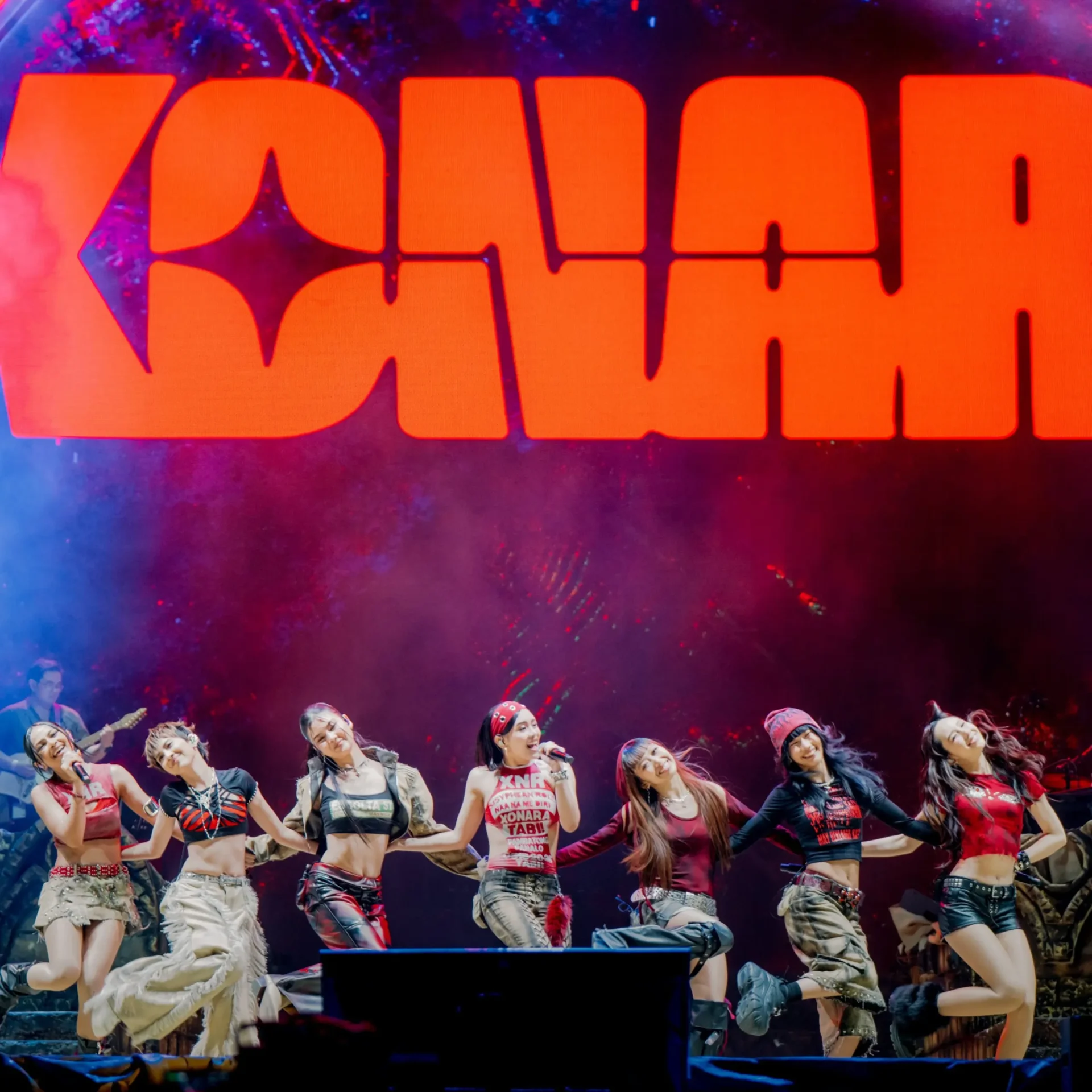 Group of young dancers performing on a lit stage in colorful outfits, arms raised in a choreographed pose with a large orange backdrop that reads 'DANCE' behind them.