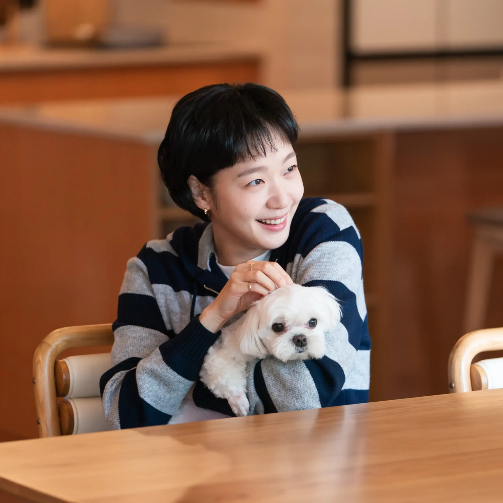 Woman with short black hair smiling while holding a small white dog at a wooden table indoors.