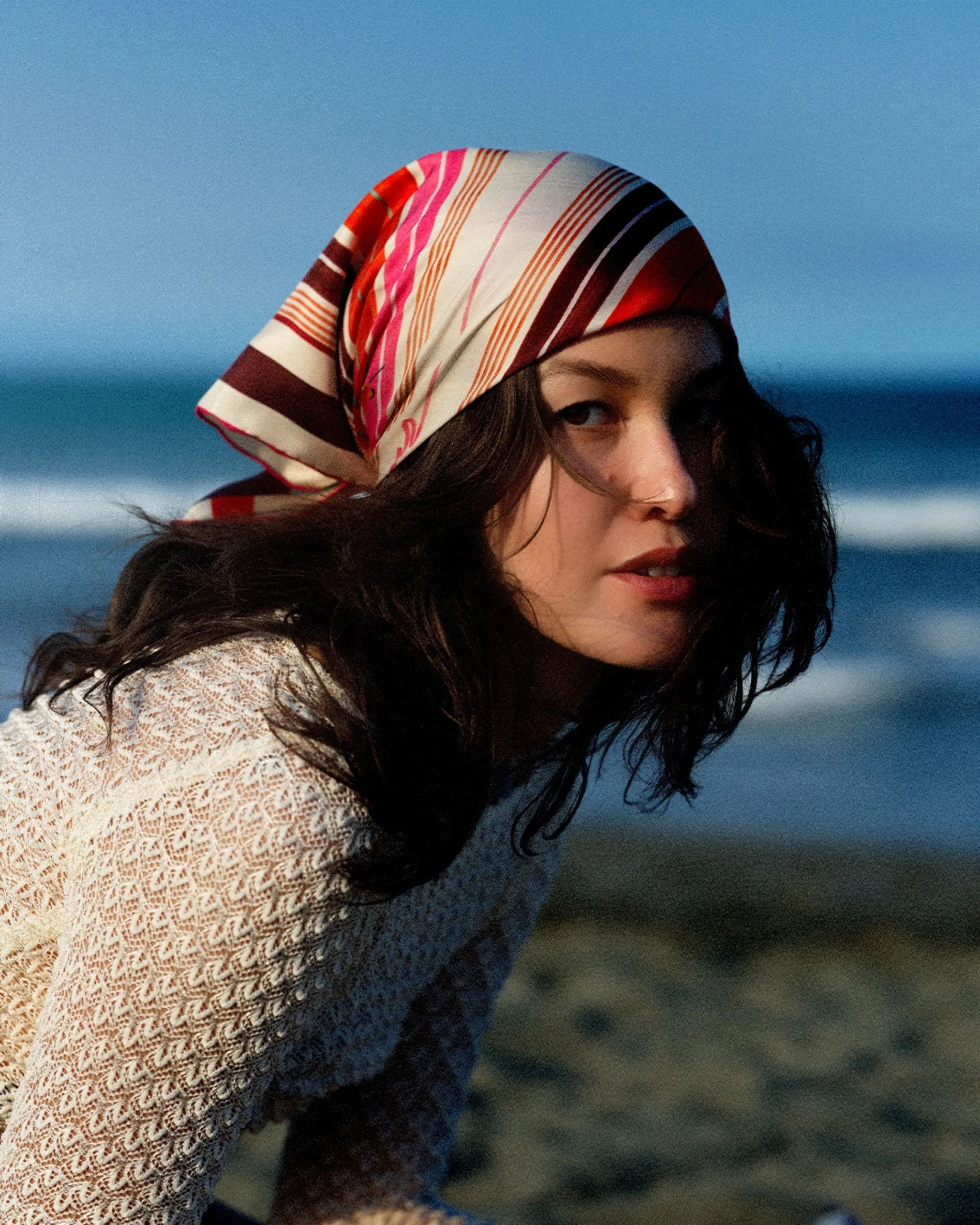 Woman on a beach wearing a colorful striped headscarf and a cream knit top, looking toward the camera with the sea in the background.