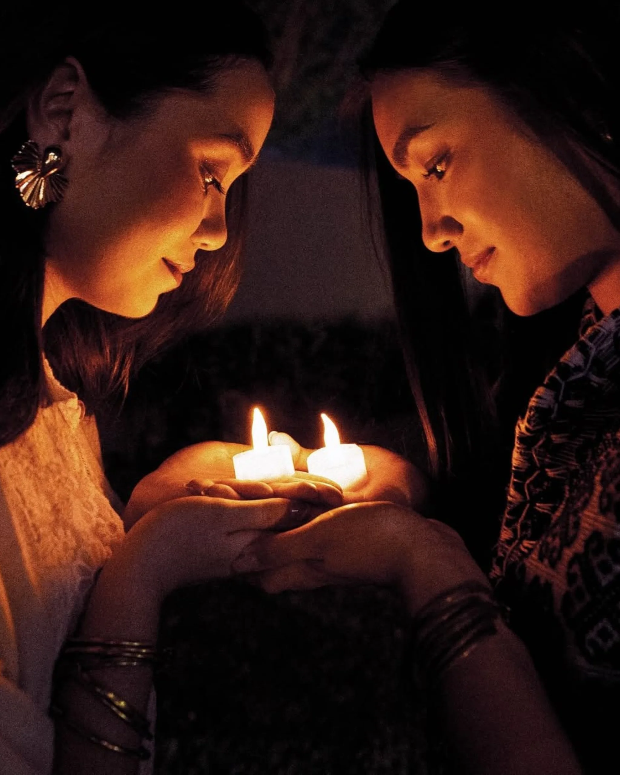 Two women hold and gaze at small lit candles, their faces bathed in warm orange light.