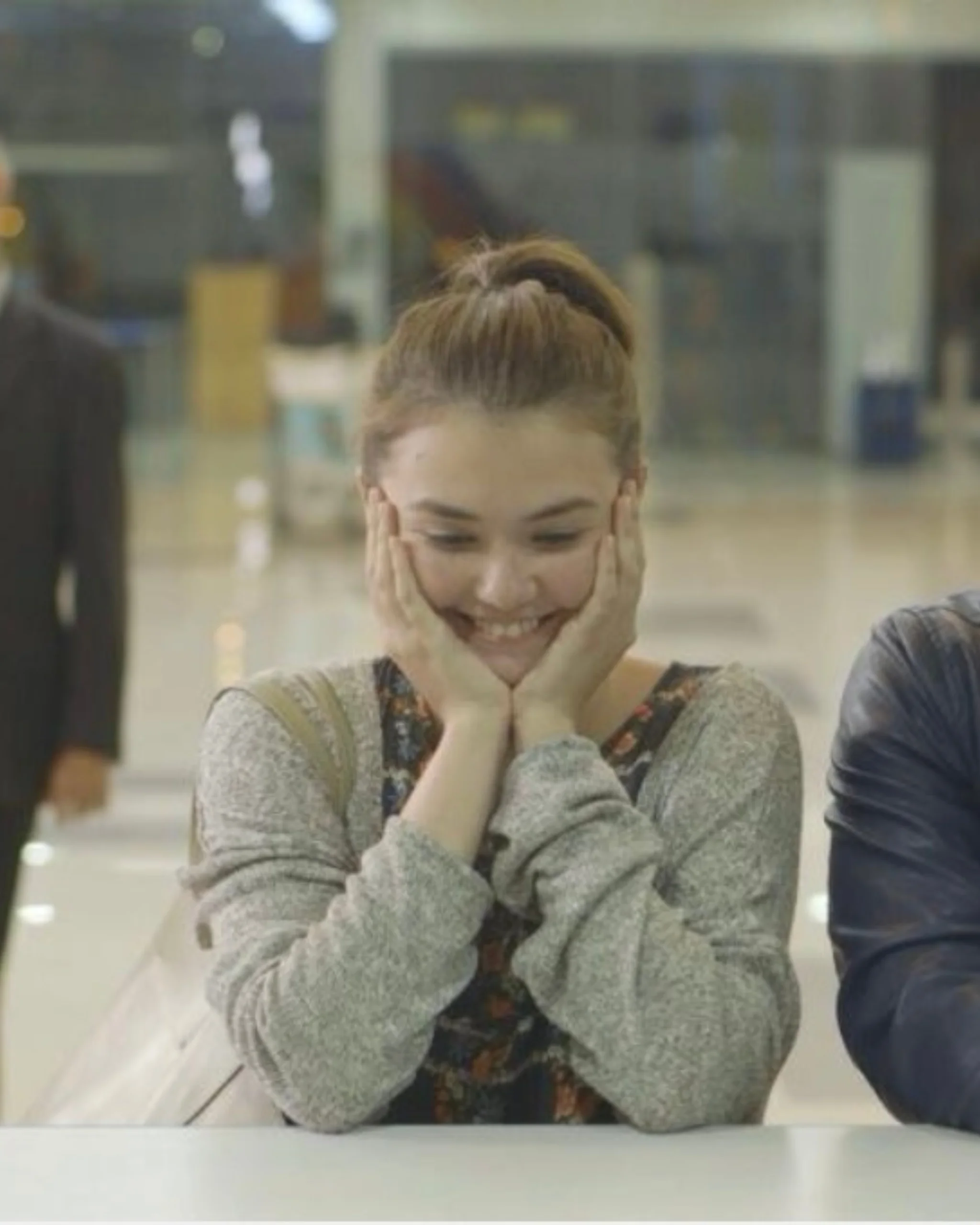Woman with her hair in a bun rests her chin on her hands and smiles softly in a bright indoor space.