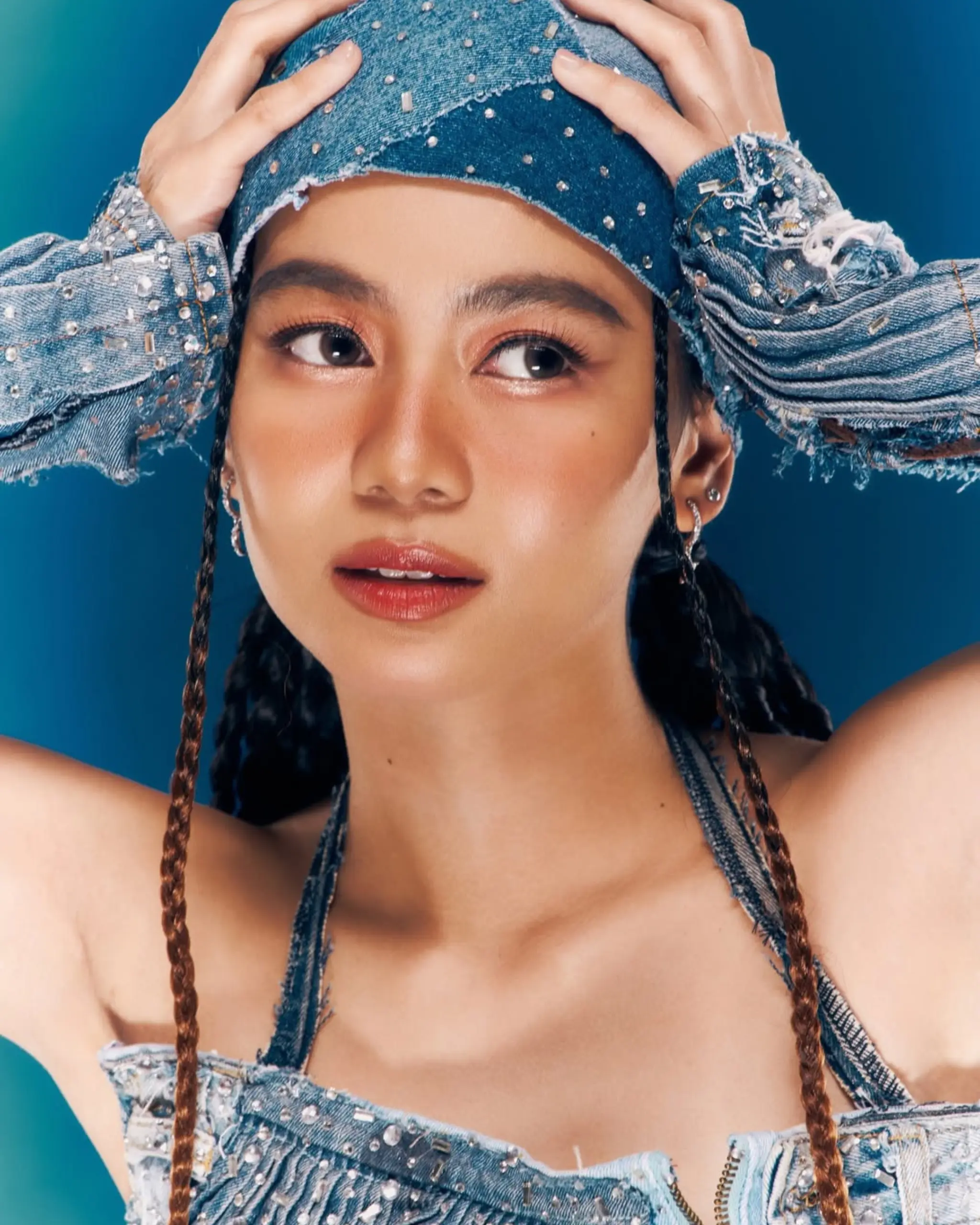 Close-up portrait of a woman wearing a beaded denim outfit, hands on her head, against a teal blue background.