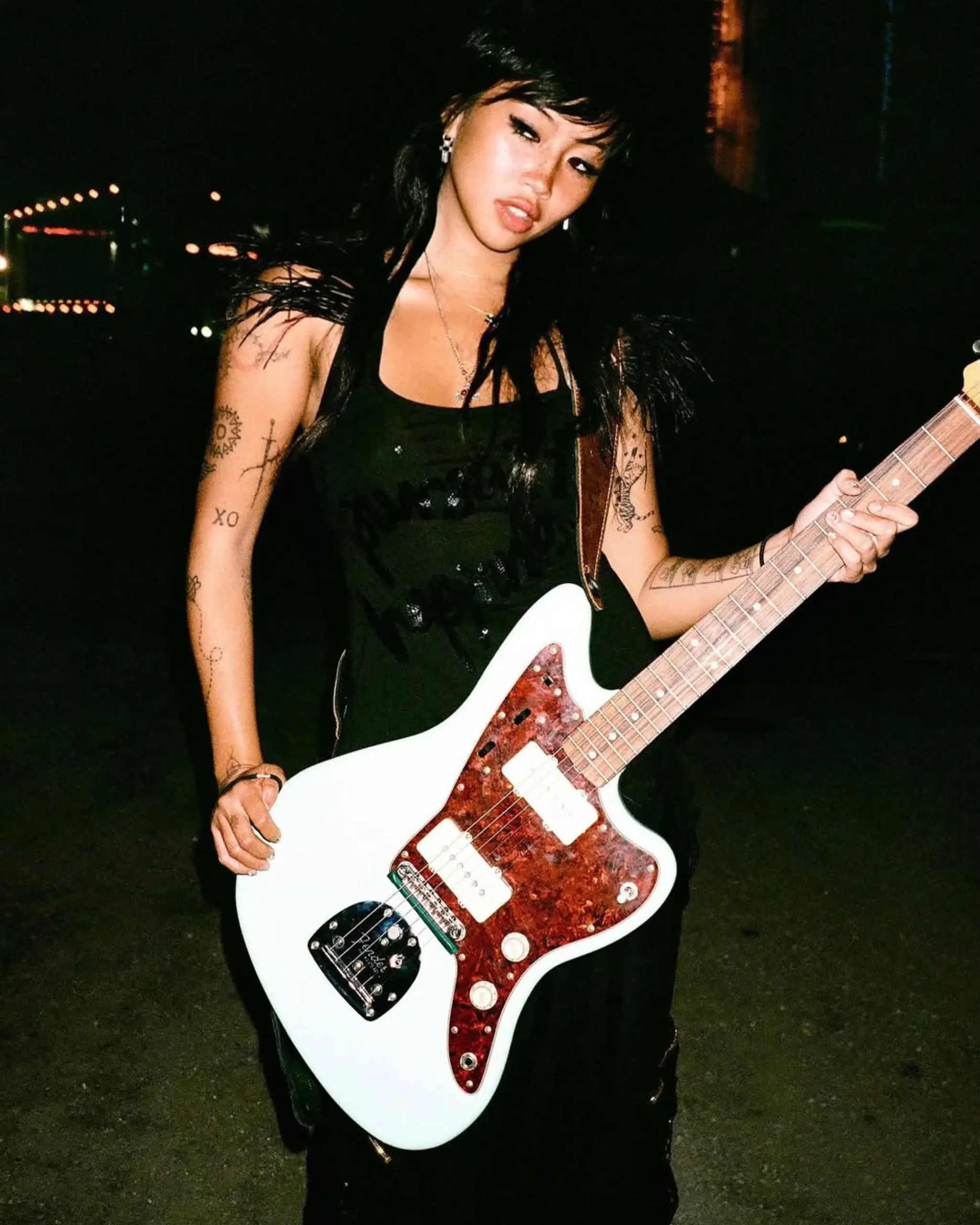 Woman at night holding a white electric guitar with a red tortoiseshell pickguard, wearing black, tattoos visible on arms and shoulders.