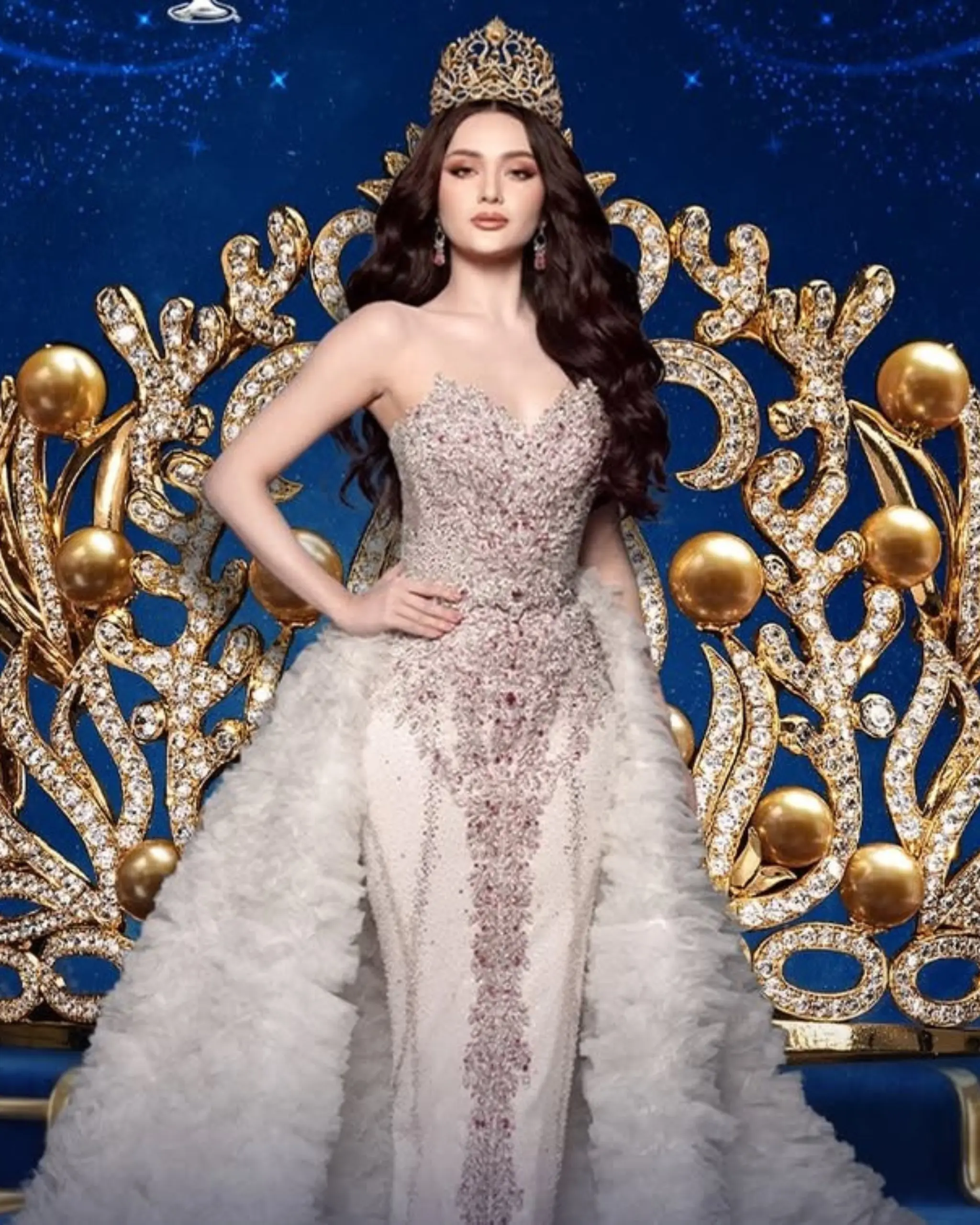 Woman in a sparkling, bejeweled gown sits on a gold, gem-encrusted throne wearing a crown against a blue backdrop.
