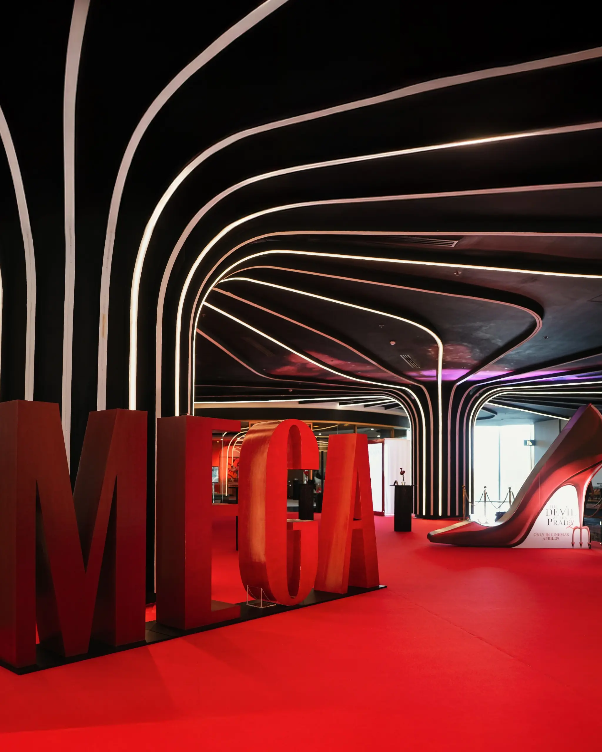Wide shot of a black interior with looping white neon arches, above a red-carpeted exhibition floor and oversized red letter sculptures at the bottom.