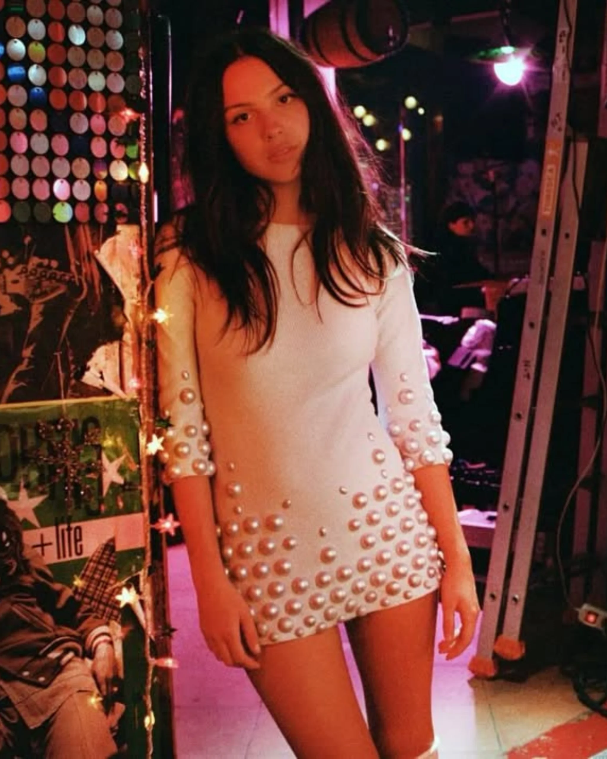 Young woman in a white pearl-embellished dress stands in a colorful backstage area with stage lights nearby, looking at the camera.
