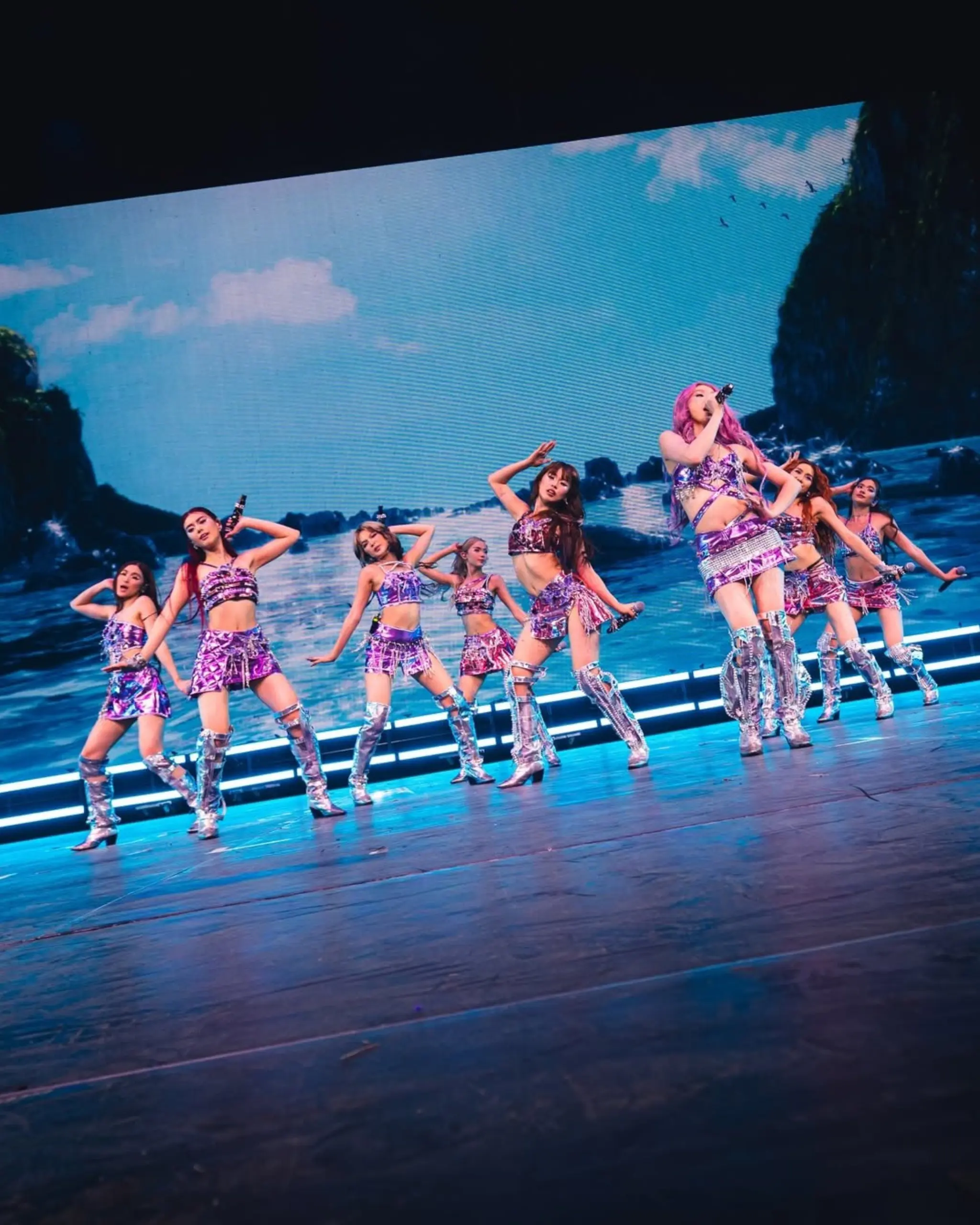 Pop group performing on a lit stage in purple outfits, with a blue ocean backdrop and a pink-haired singer at the center.