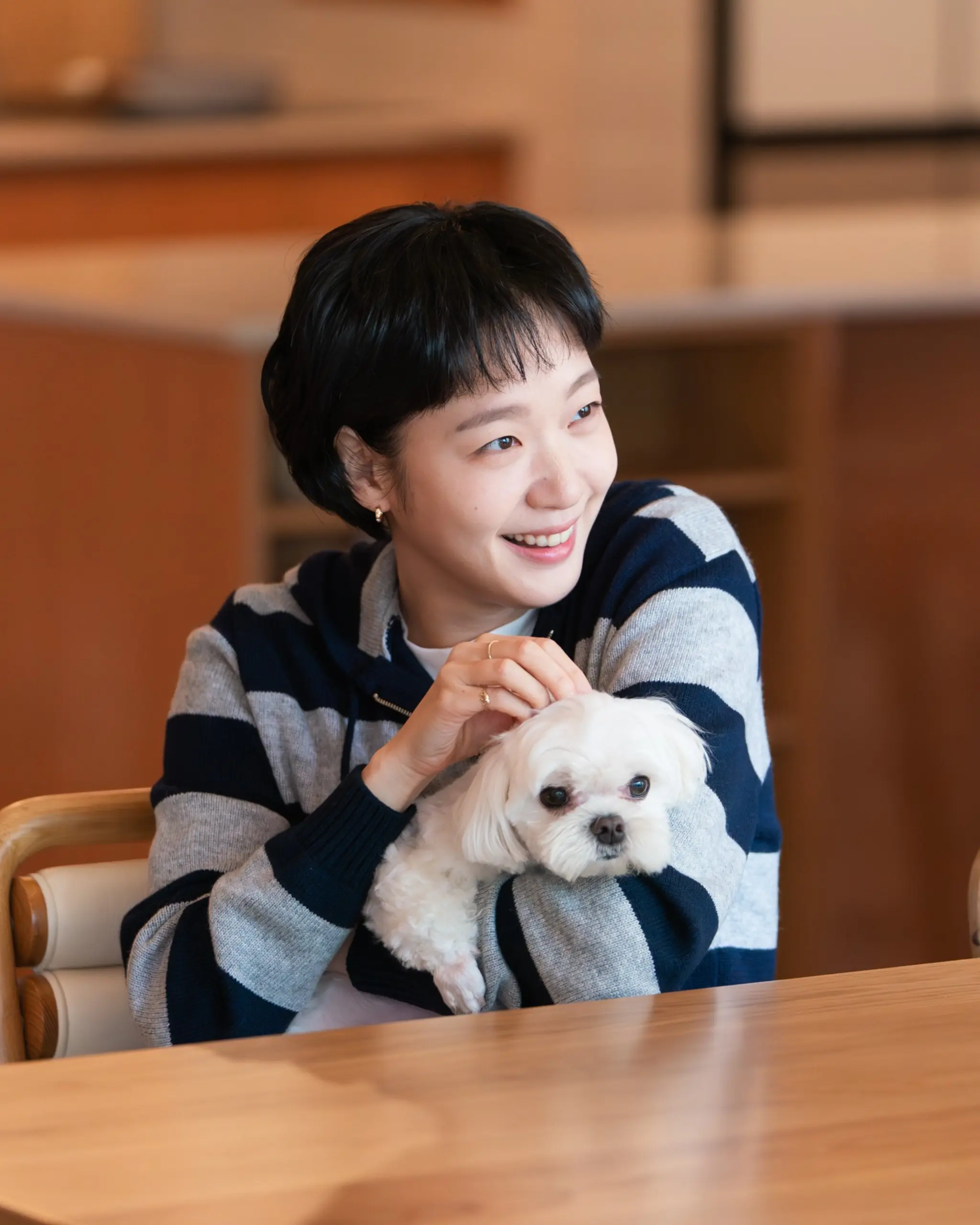 Woman smiling while holding a small white fluffy dog on her lap at a wooden table indoors