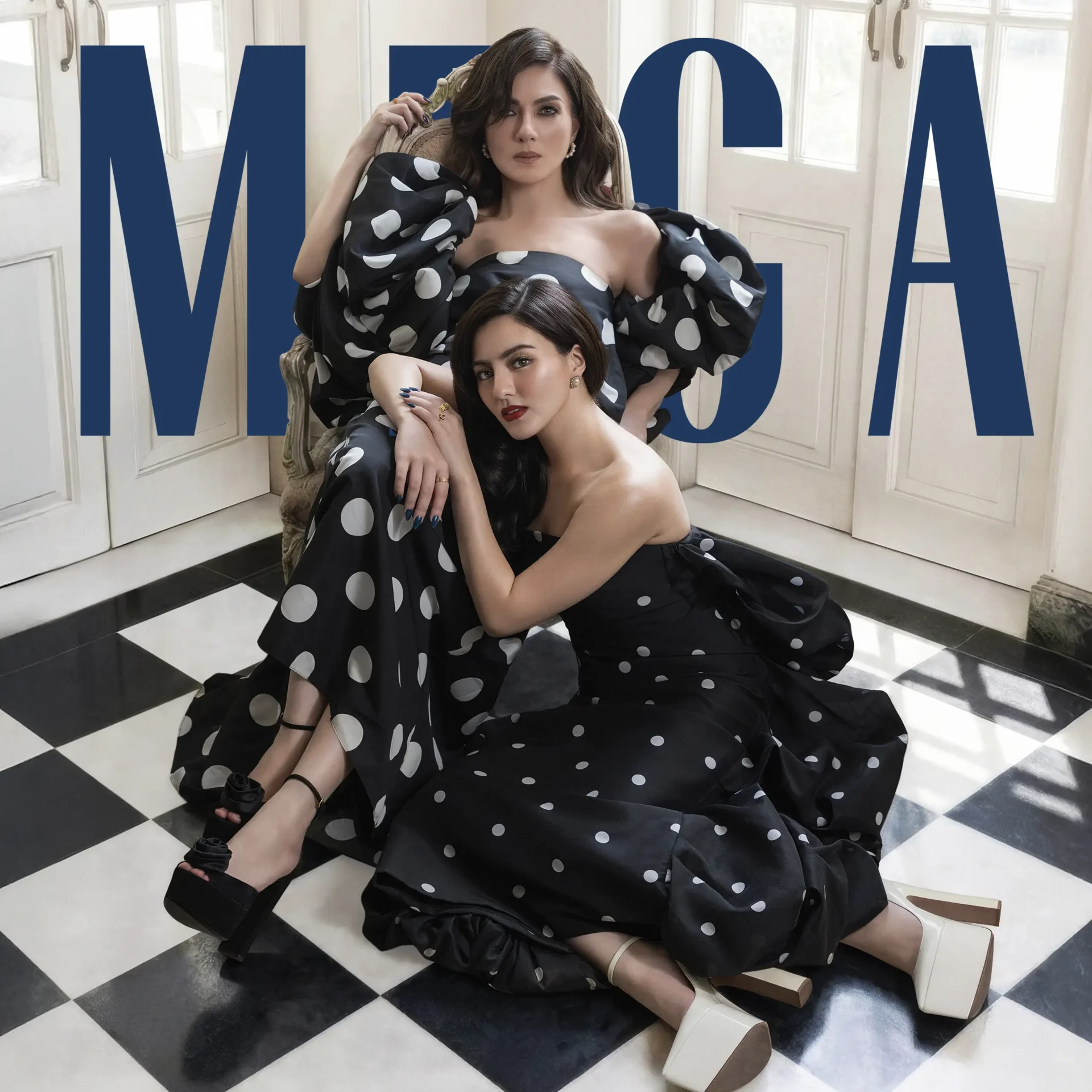 Two models in black polka-dot dresses pose on a checkered floor with oversize blue letters in the background.