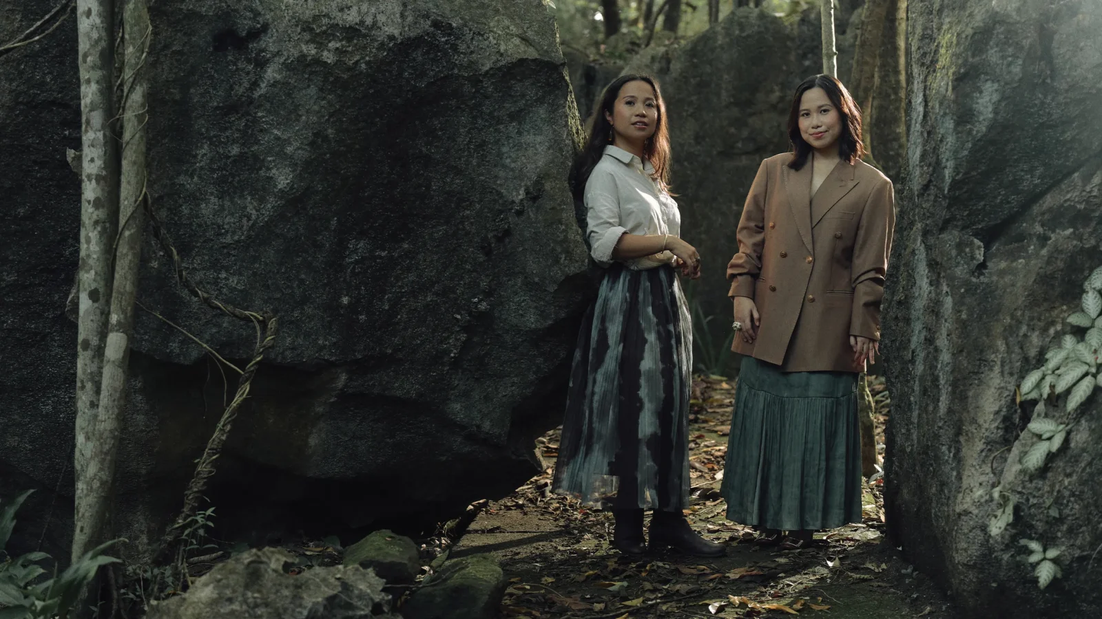 Two women stand among large rock formations in a forest, wearing long skirts and blazers.