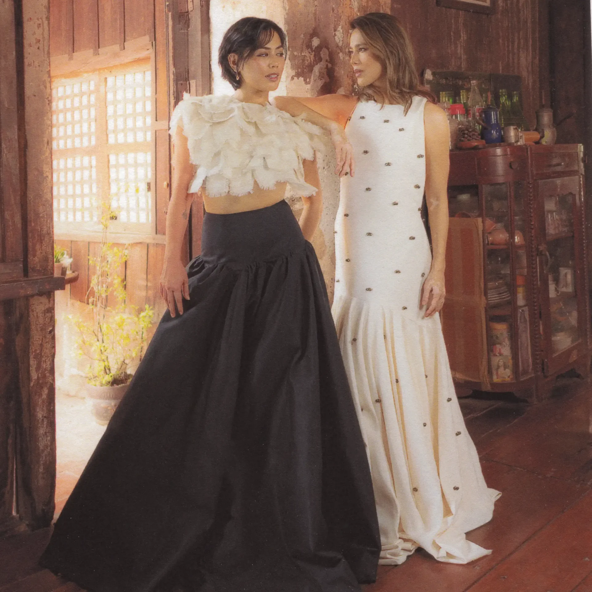 Two women in elegant dresses stand close in a rustic wooden room, one in a white ruffled top and black skirt, the other in a white buttoned gown, looking at each other.