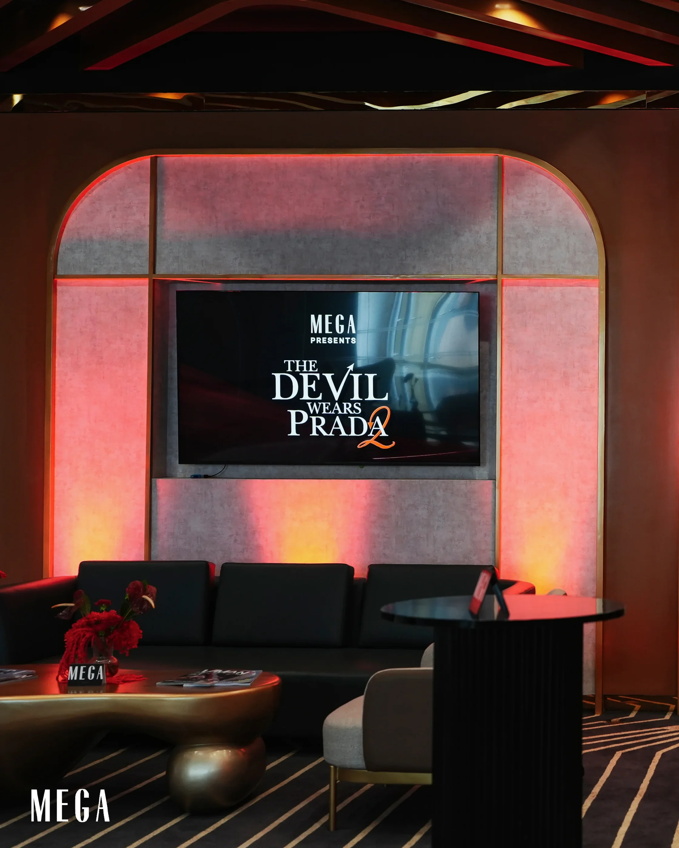 Lounge scene with pink backlit arch panels and a wall-mounted screen reading 'MEGA PRESENTS THE DEVIL WEARS PRADA 2'; foreground shows a table with red flowers and seating nearby. 2026-05-01