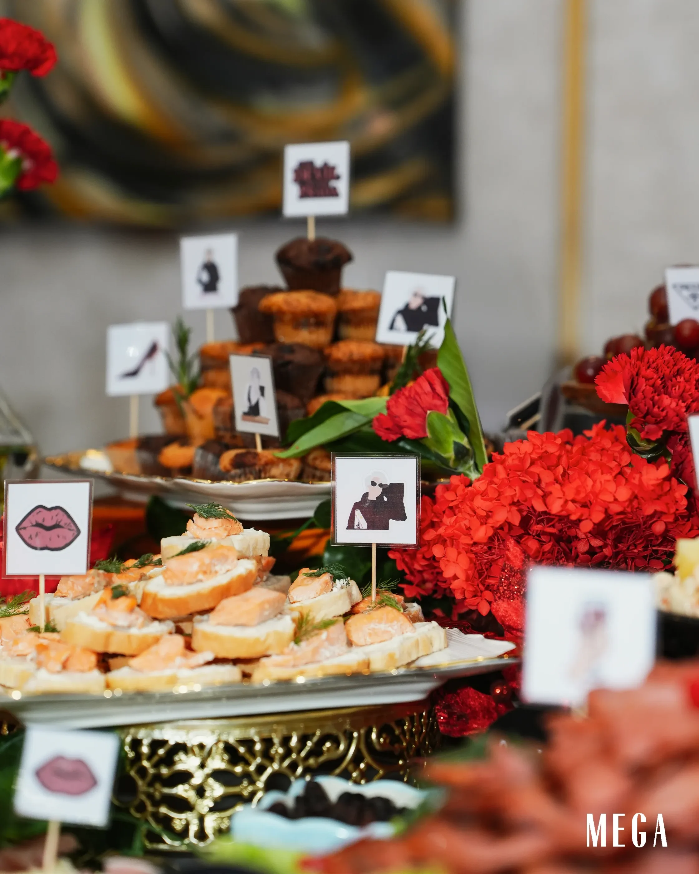 Assorted bite-sized pastries with small signs on sticks, arranged on a dessert table with red flowers nearby.