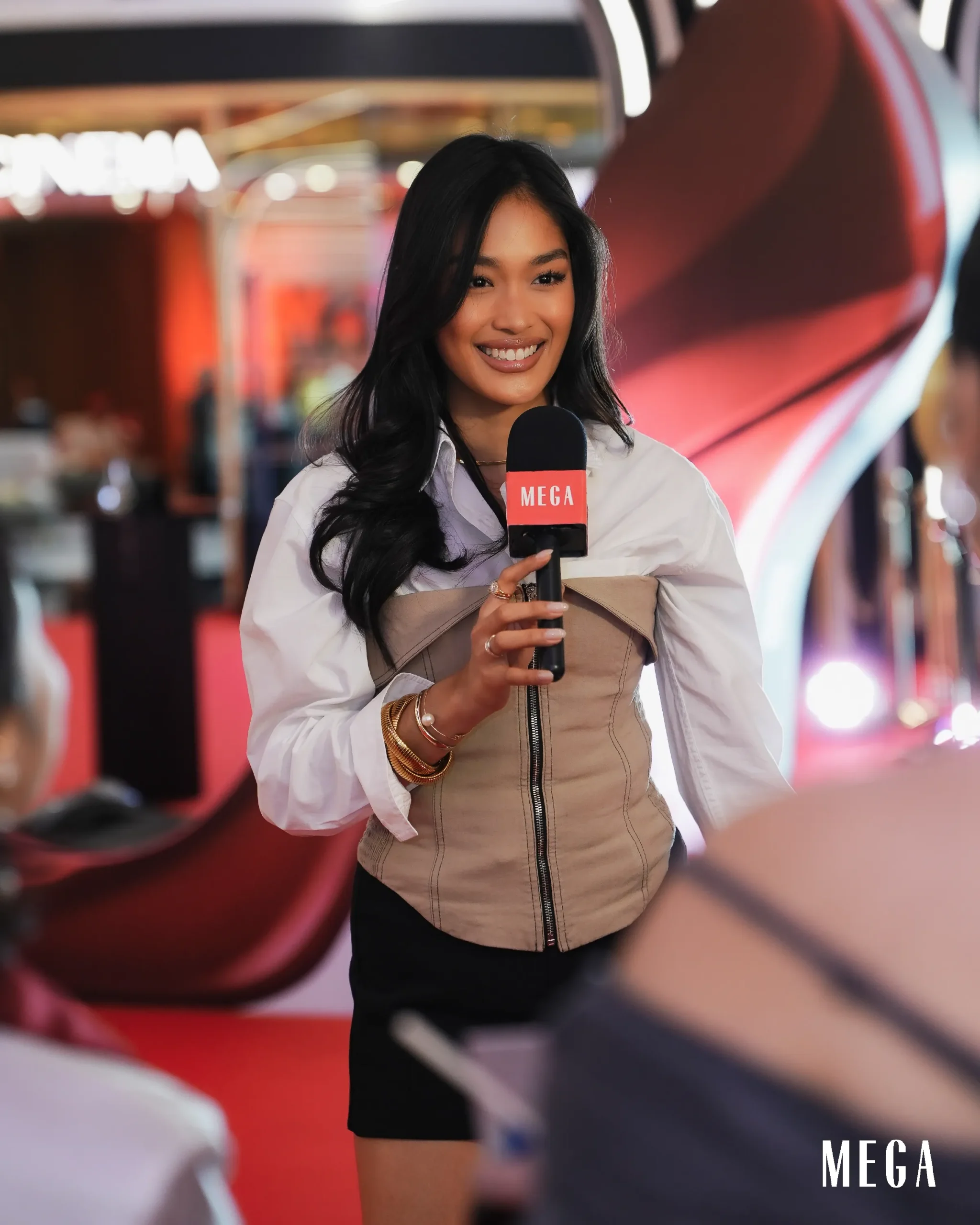 Smiling female reporter holding a MEGA-branded microphone during a live event in a red-themed backdrop.