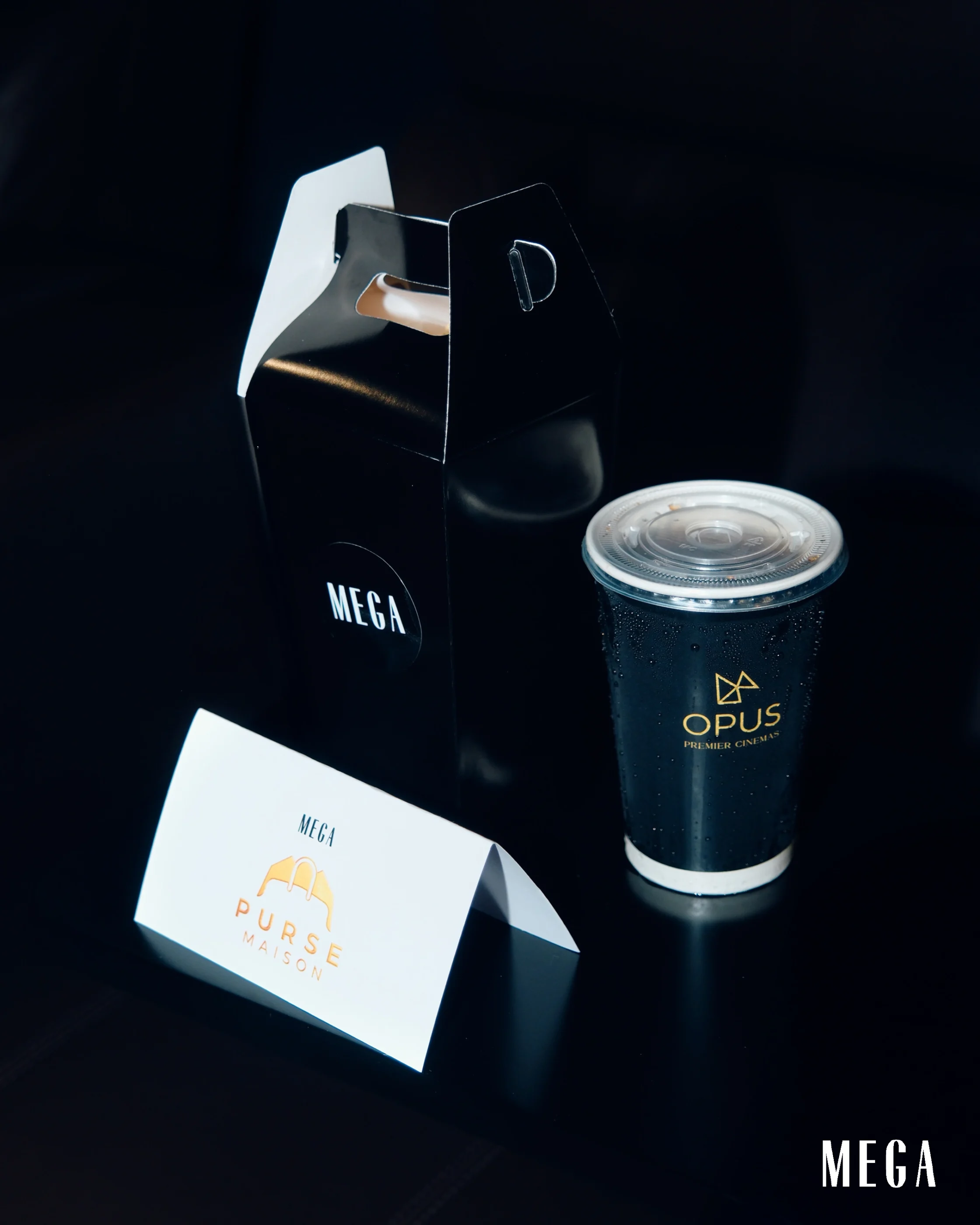 Close-up of a glossy black gift box with an open flap, a black can with an Opus logo beside it, against a dark background, with a white card labeled MEGA in the foreground.