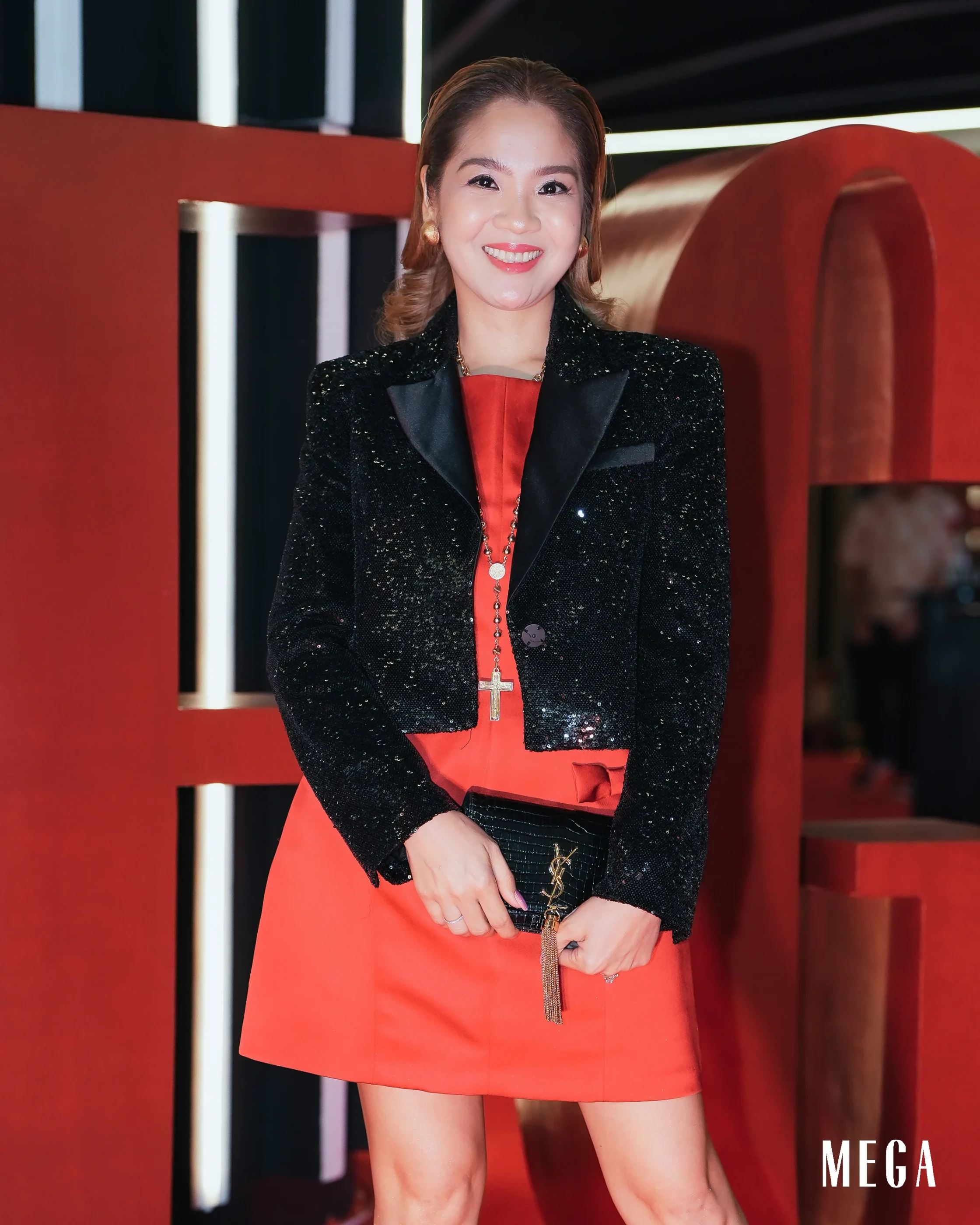 Smiling woman in a black sequin blazer over a red dress, posing at a red backdrop with MEGA logo visible.