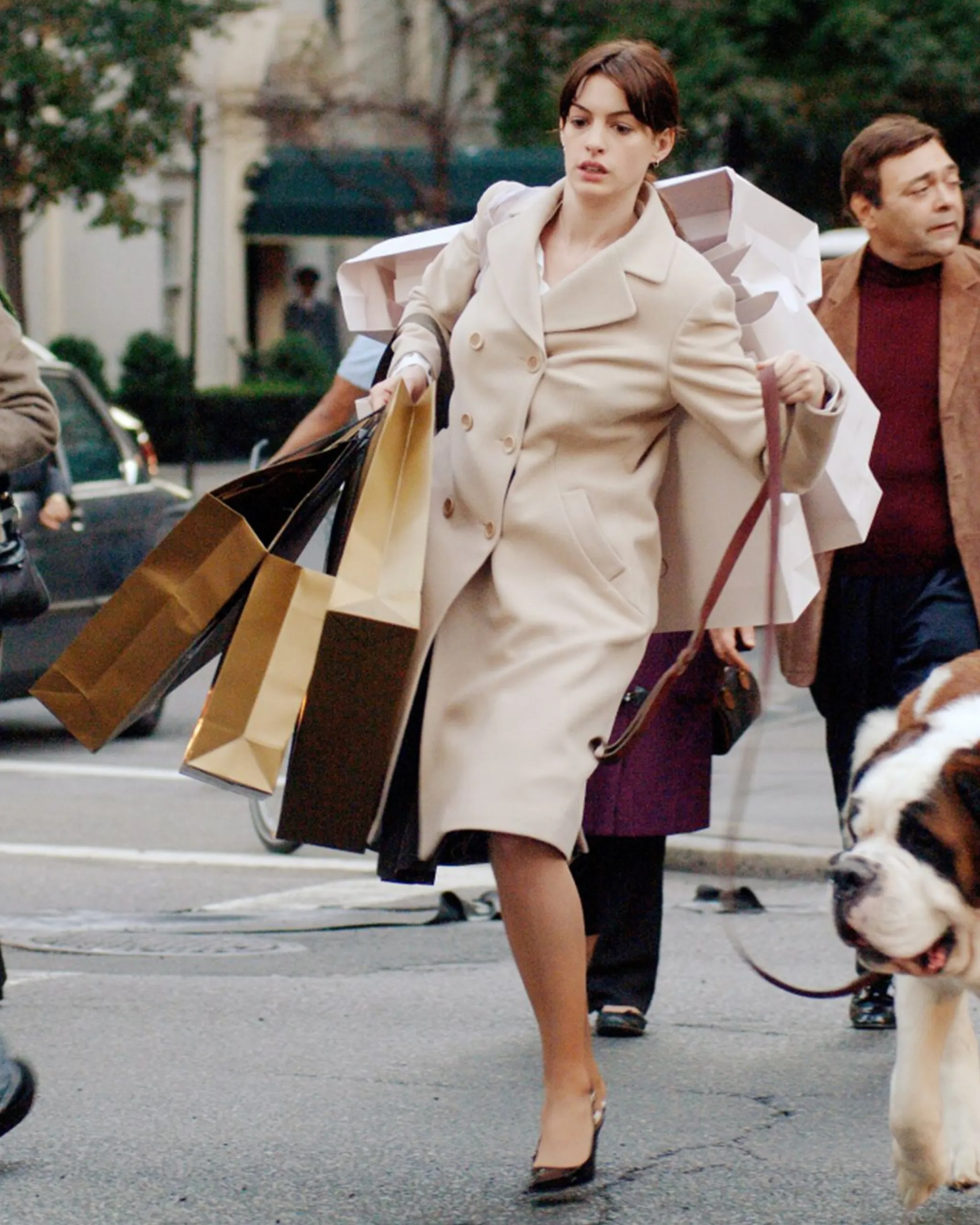 Woman in a beige trench coat carries multiple shopping bags while walking on a city sidewalk, with a dog nearby on a leash.