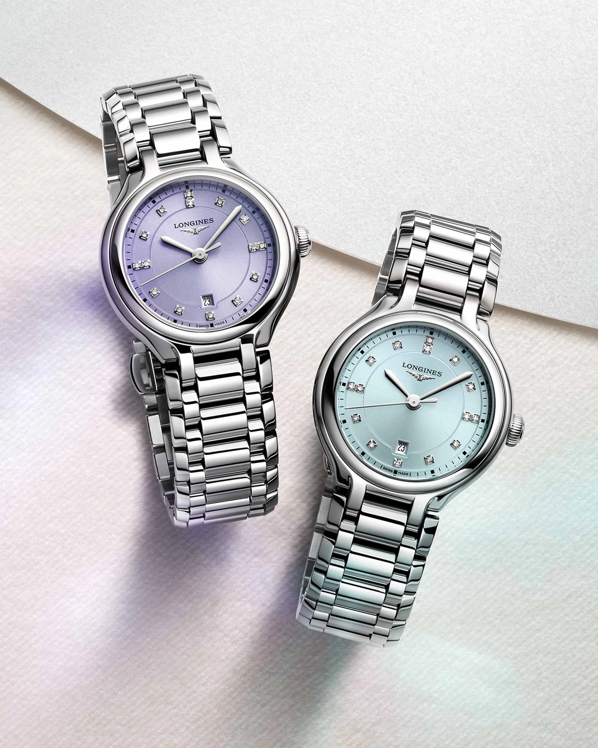 Two Longines women's watches with silver metal bracelets, lavender dial on the left and light blue dial on the right, on a soft pastel background.