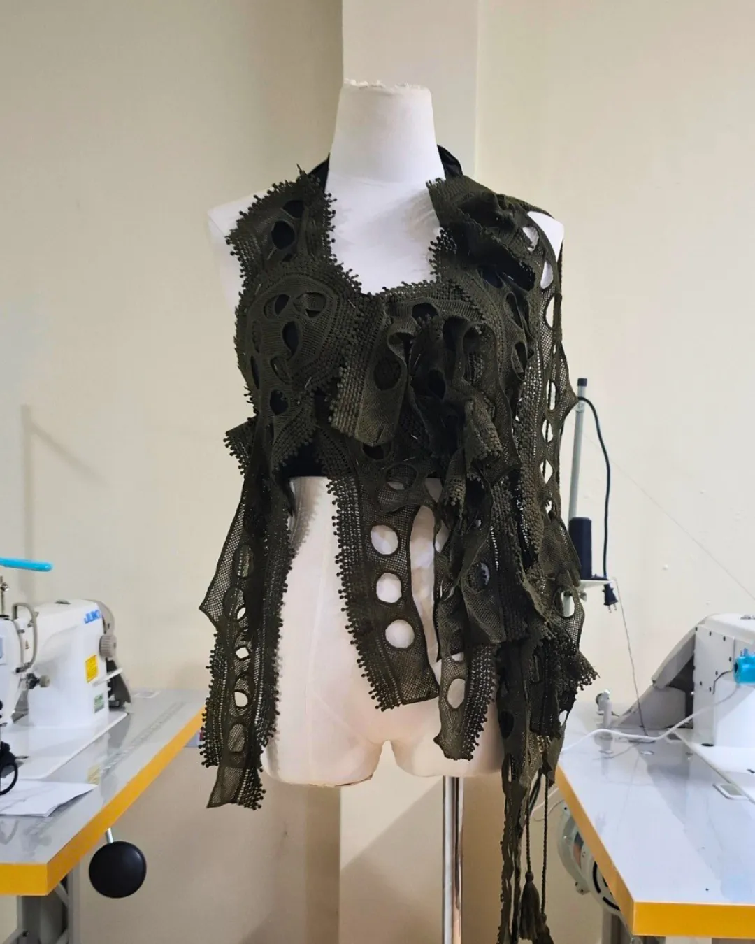 Avant-garde olive-green knitted garment with circular cutouts on a dress form in a sewing studio.