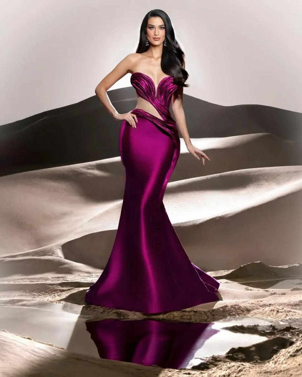 Woman in a strapless magenta gown with cutouts standing on desert dunes, with her reflection visible in a shallow pool of water.